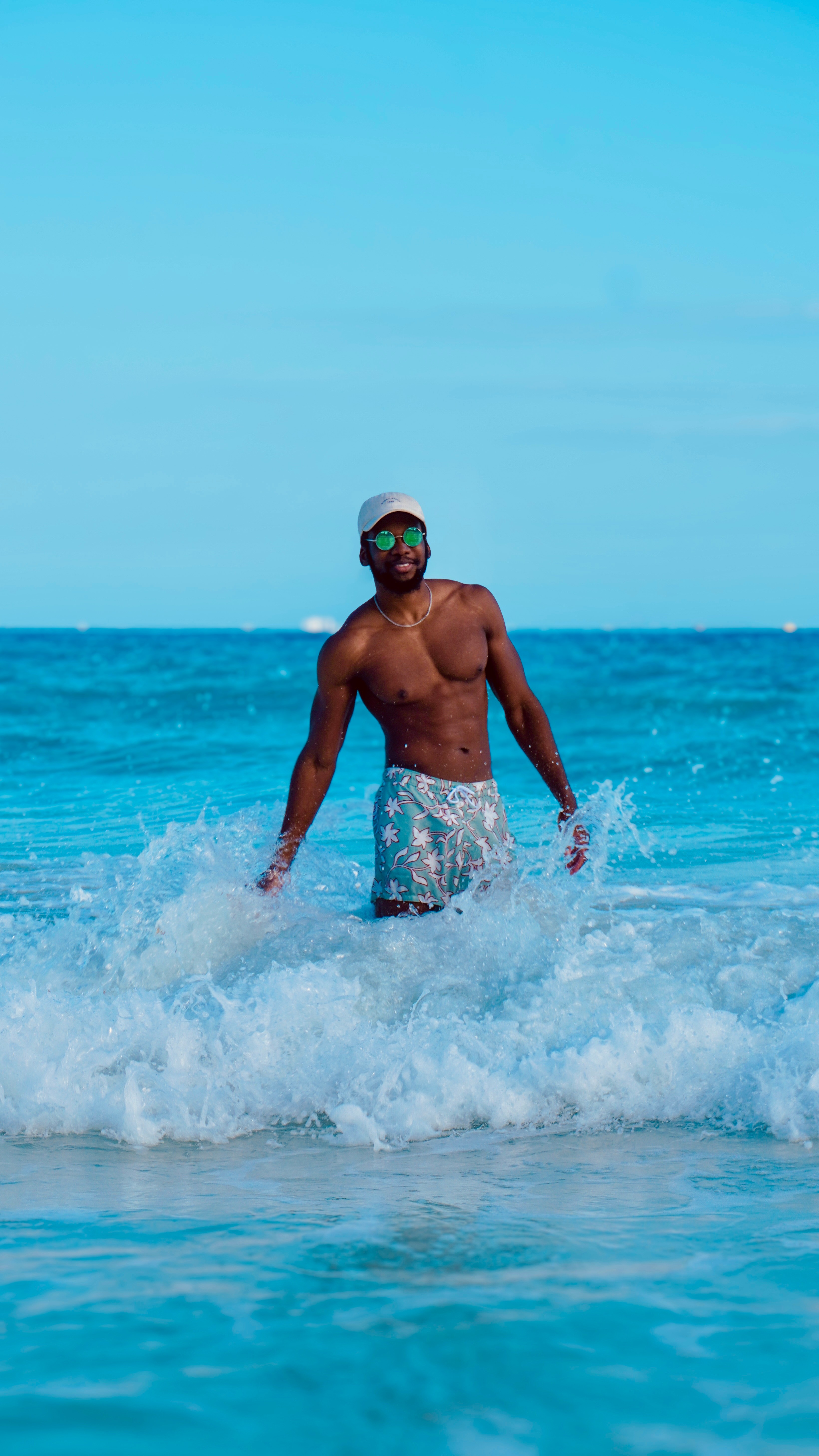 Man standing in ocean waves, wearing sunglasses and swim trunks, with a clear blue sky and sea in background.