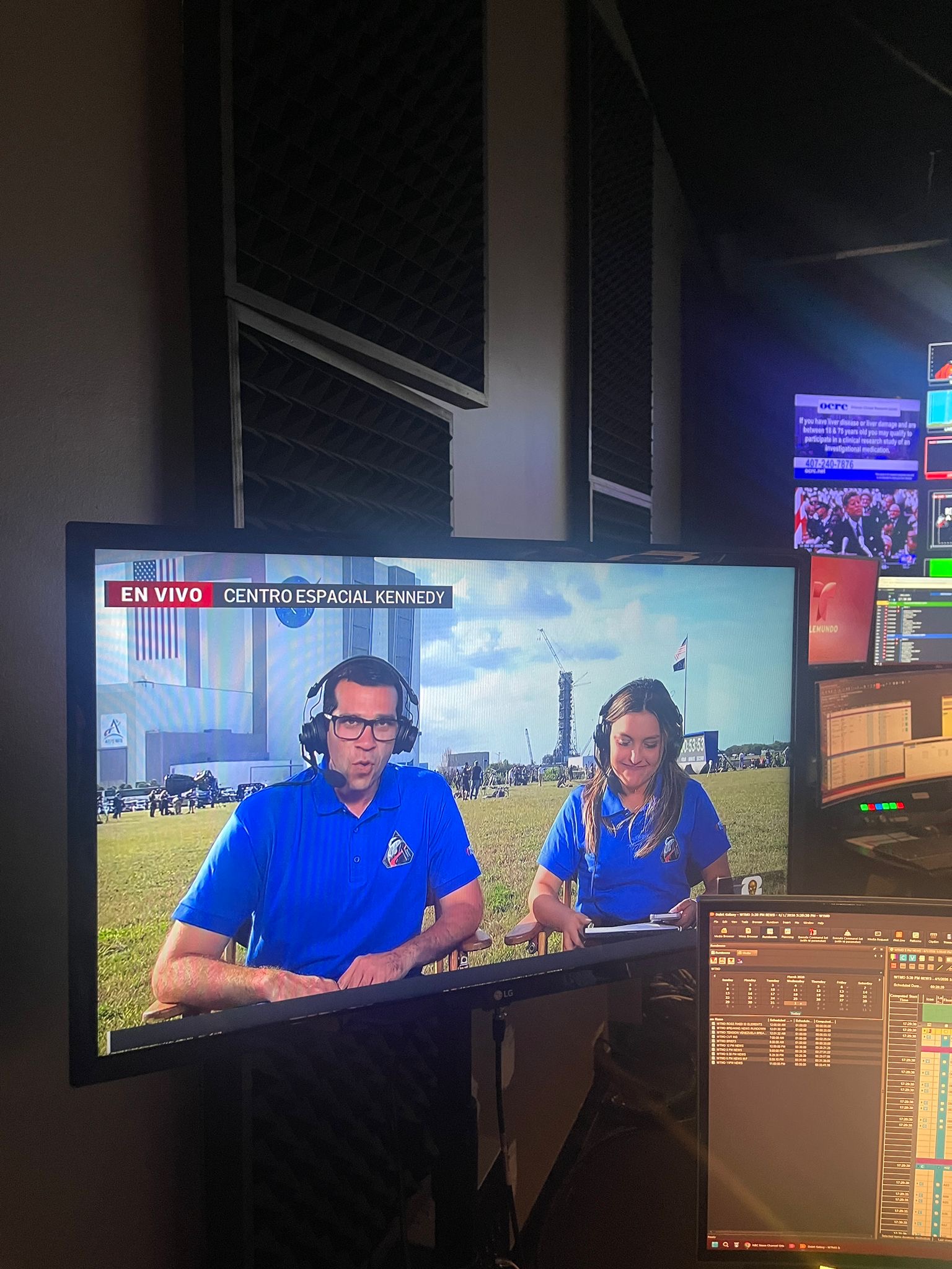 Two news anchors sit at a desk on a television set with a cityscape background, wearing blue shirts, during a live broadcast.