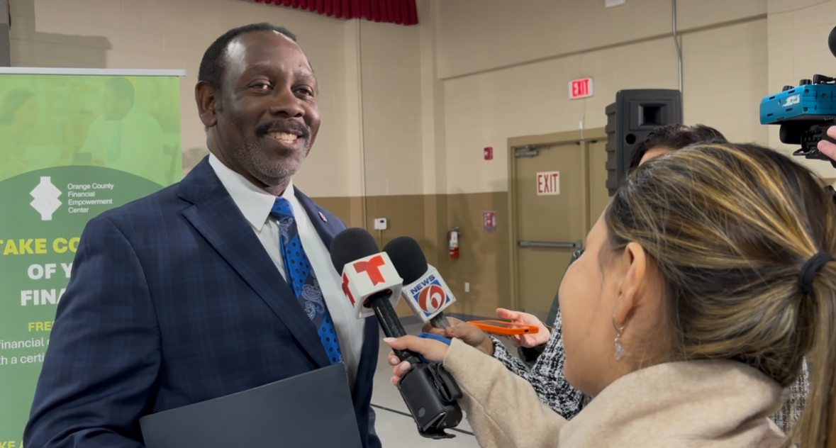 Man in a suit being interviewed by reporters in a room with a green banner, microphones, and cameras.
