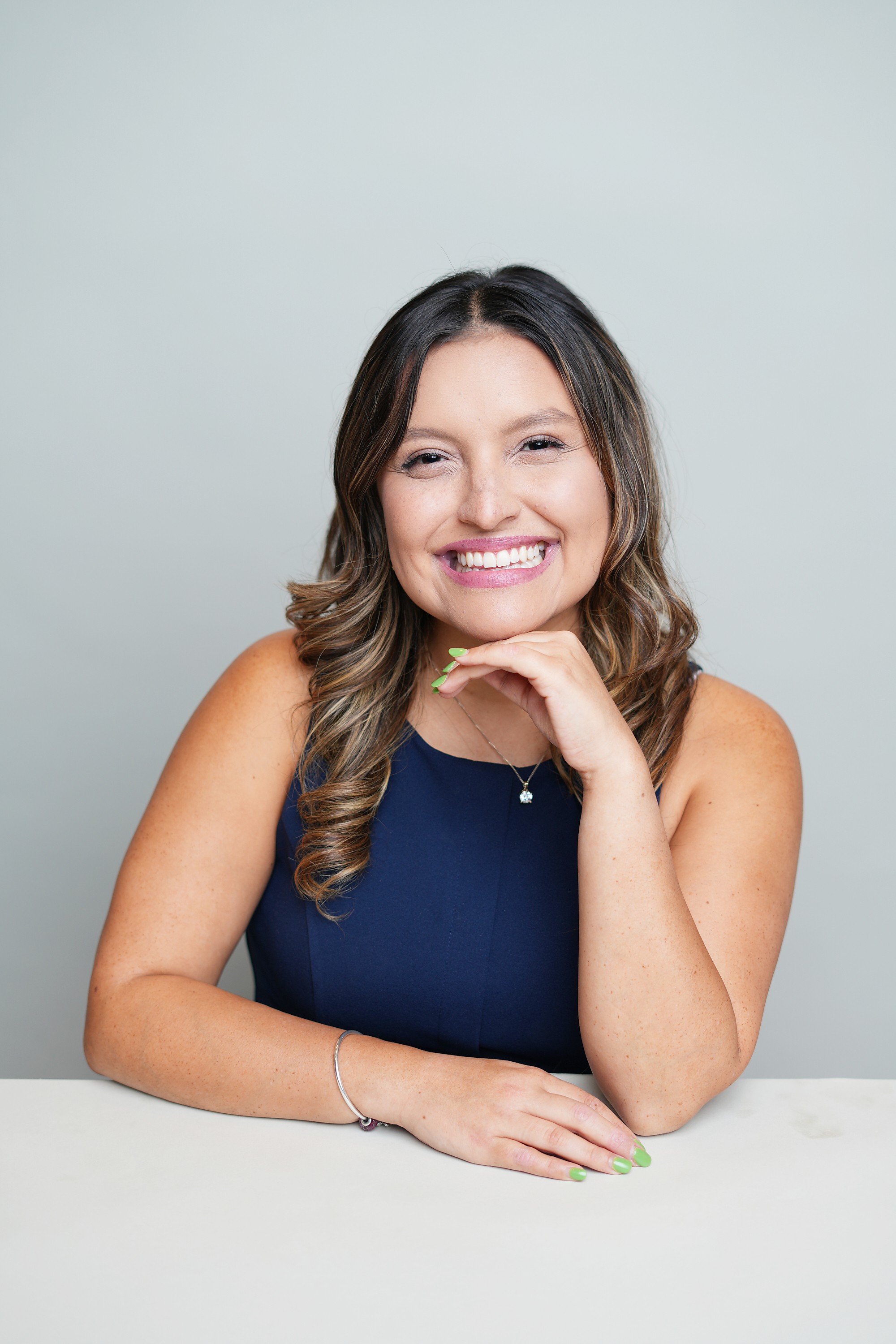 Smiling woman with wavy hair resting her chin on her hand, wearing a dark sleeveless top, against a plain light background.
