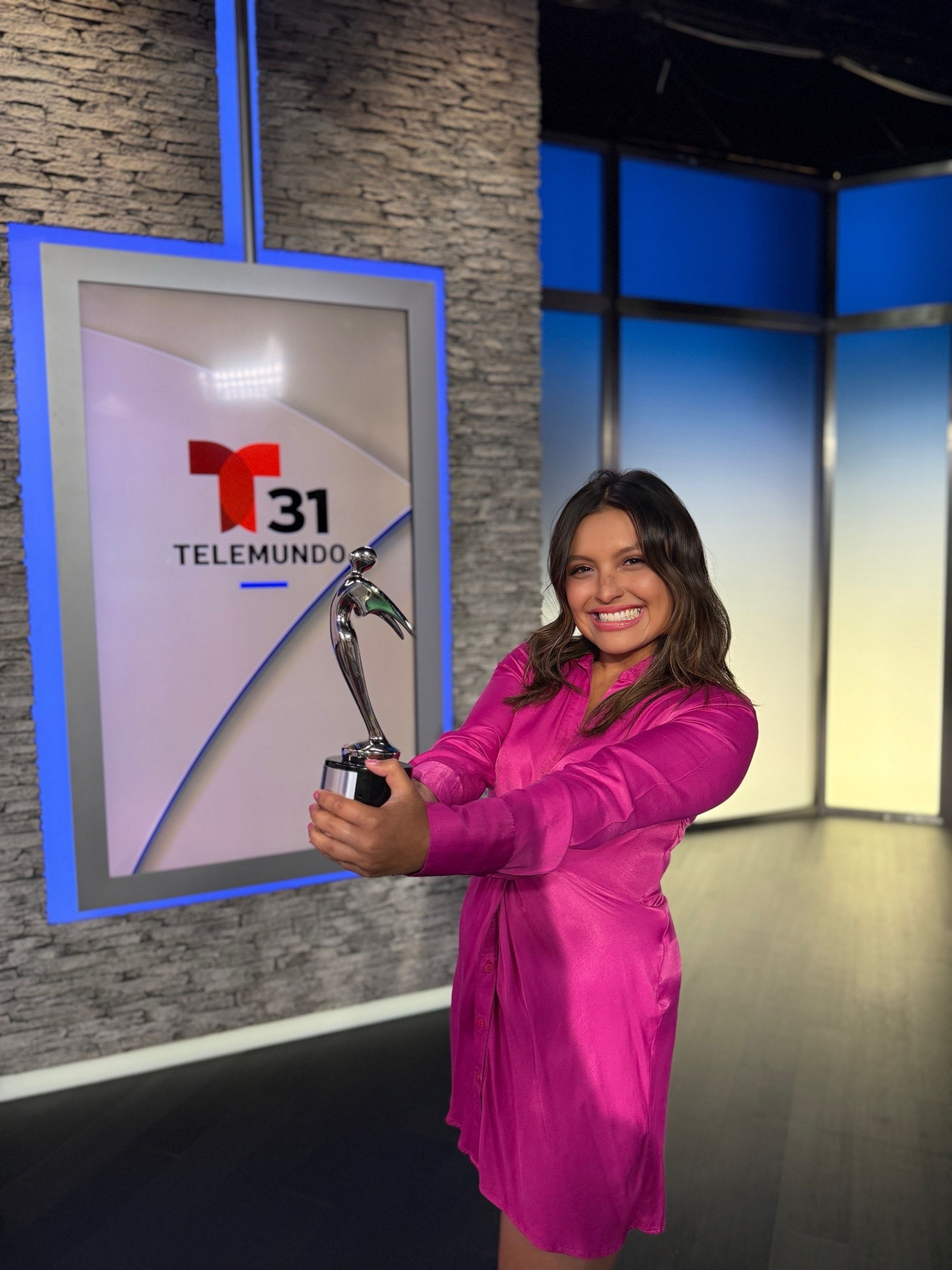 Woman in pink dress holding a trophy in a TV studio with Telemundo logo on screen.