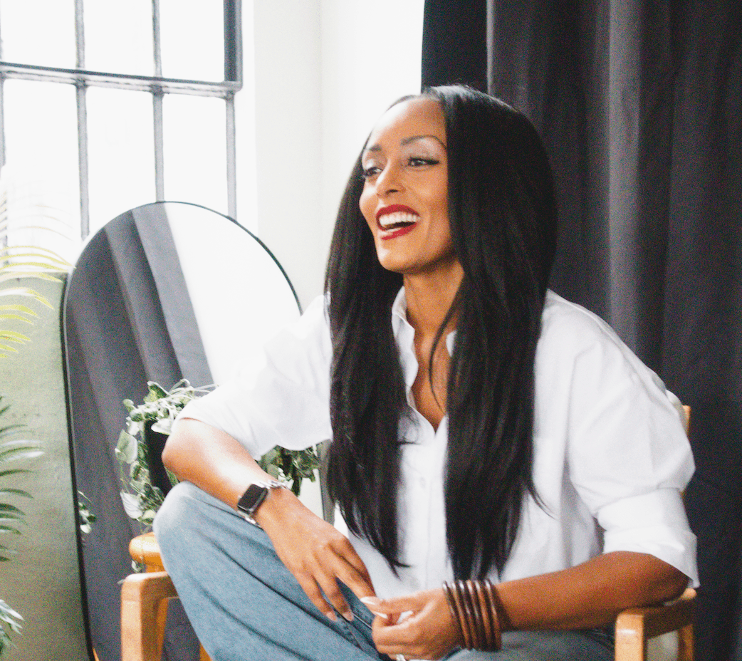 Woman with long black hair, red lipstick, white shirt, sitting indoors near a window and mirror, smiling.