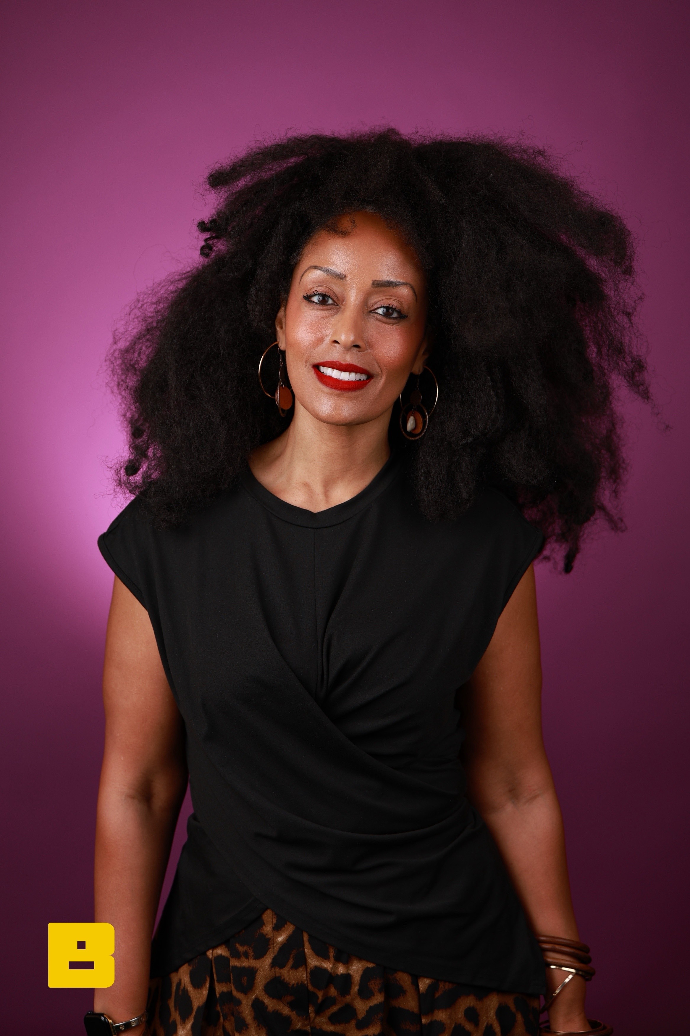 Woman with voluminous curly hair, wearing a black top and leopard print skirt, smiling against a purple background.