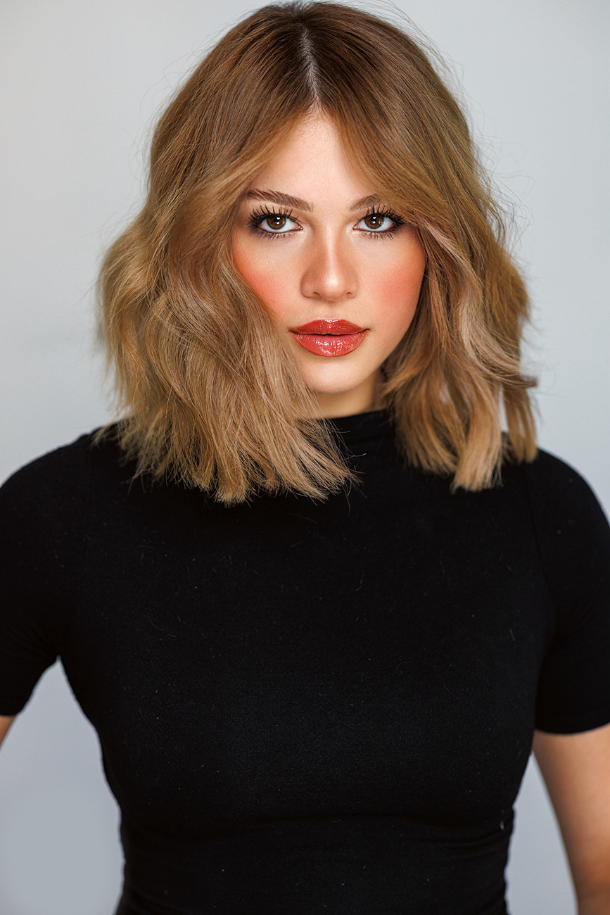 Woman with shoulder-length wavy hair, wearing a black top, looking at camera with neutral background.