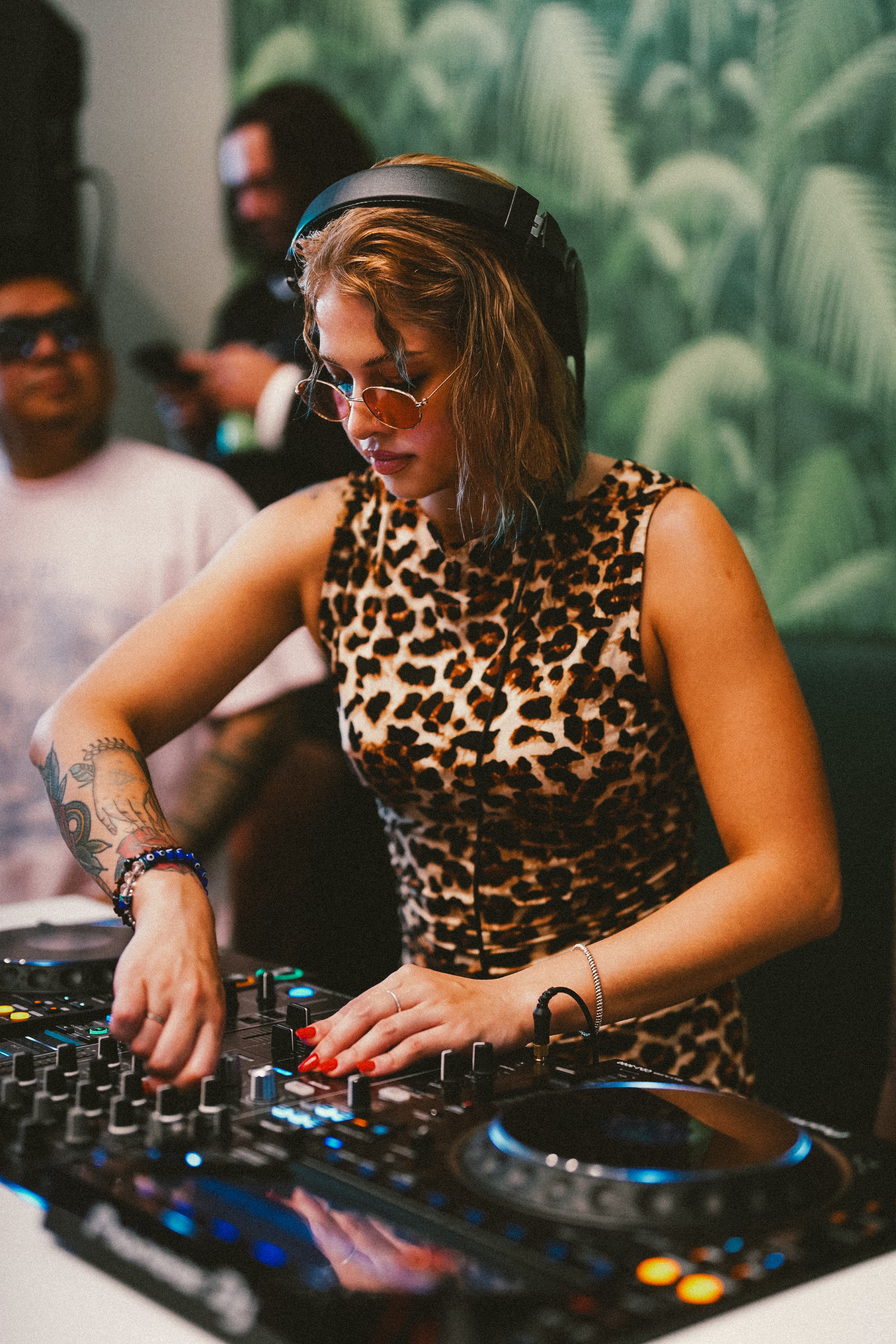 Female DJ with headphones and sunglasses, wearing a leopard print top, operating DJ equipment in a club setting.