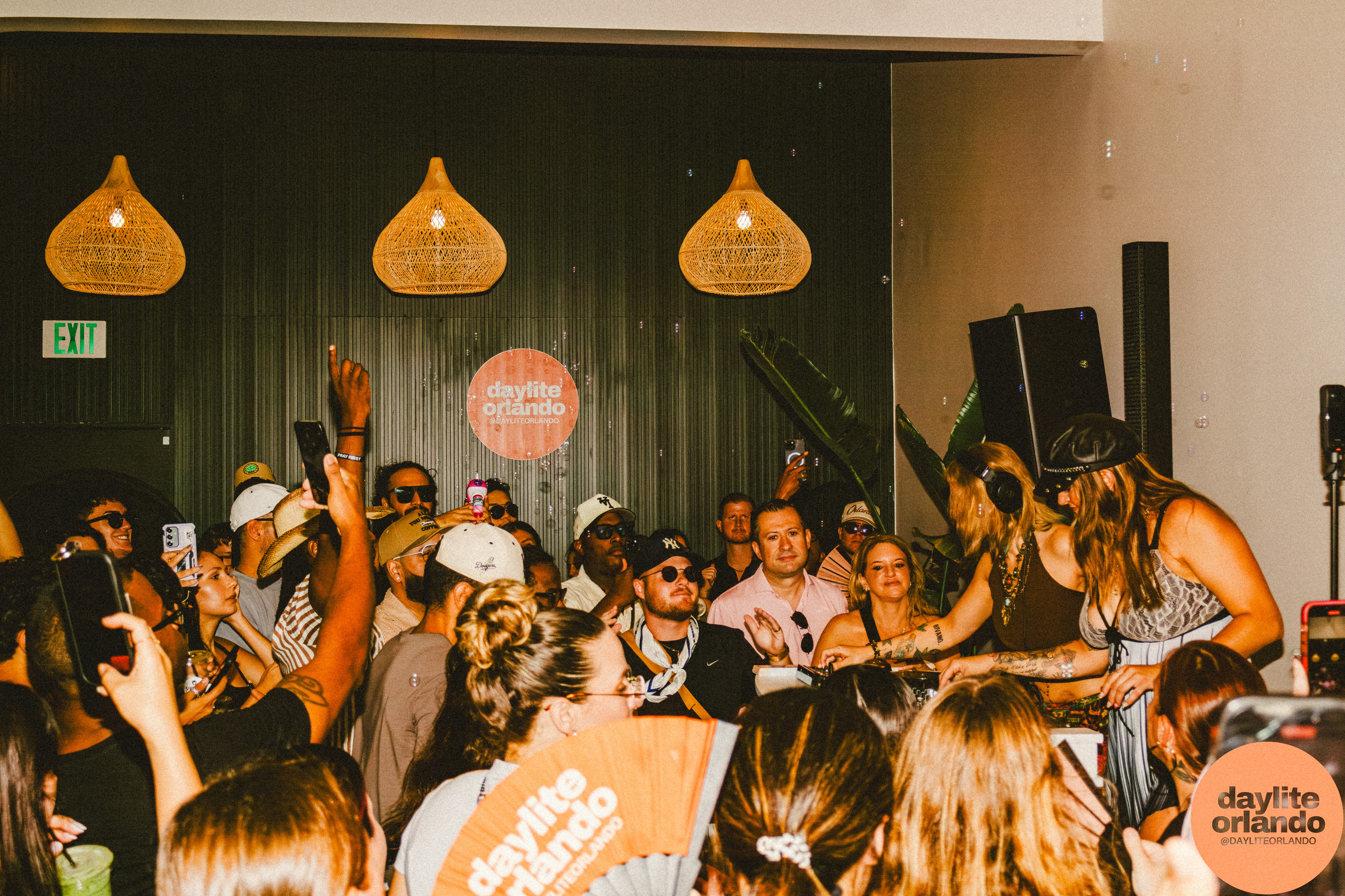 Crowd of people at a lively indoor event with hanging lights and a DJ, some raising hands and taking photos.