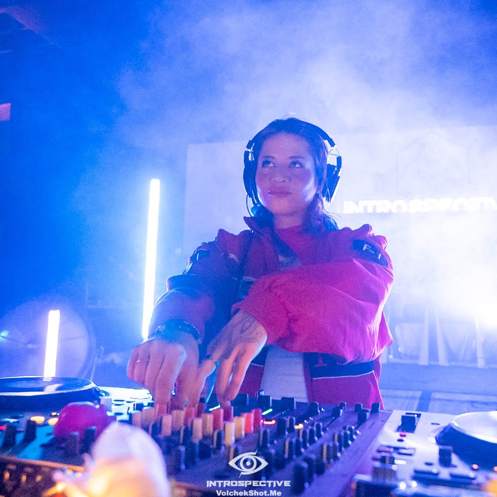 Female DJ wearing headphones, standing behind a mixing console with colorful controls, in a vibrant club setting with blue lighting.