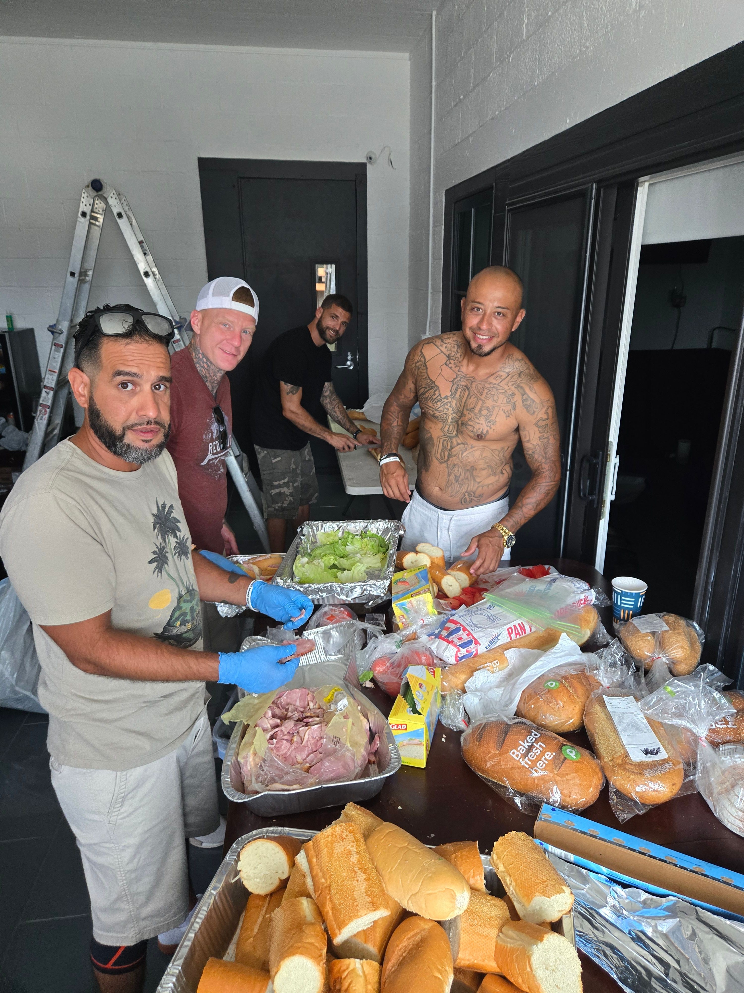 Three men are preparing food and bread on a table with various bread and meat items, indoors.