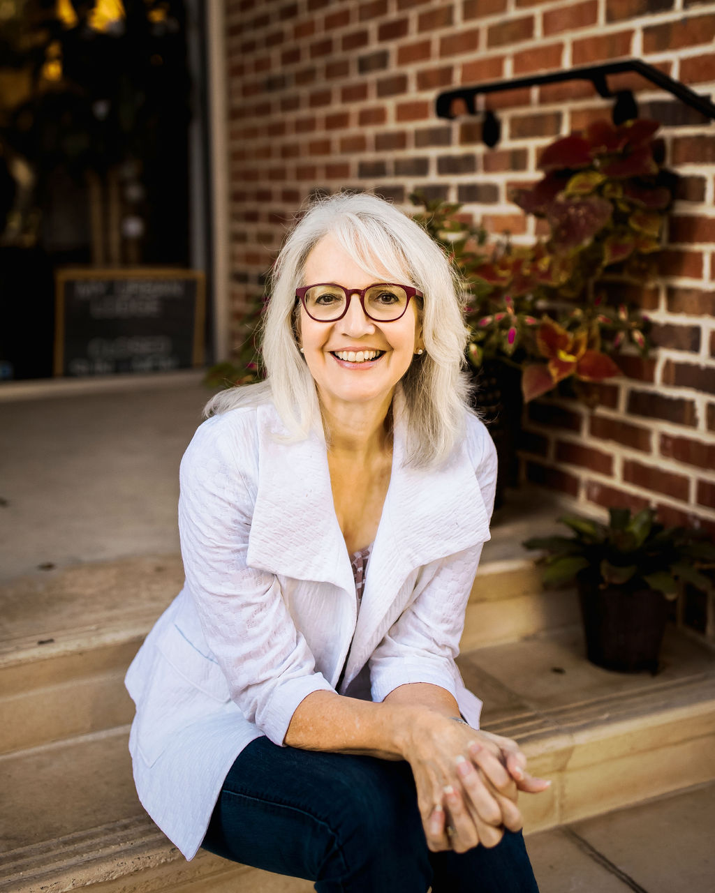 Smiling elderly woman with glasses and white hair sitting outdoors near a brick wall, wearing a white jacket.