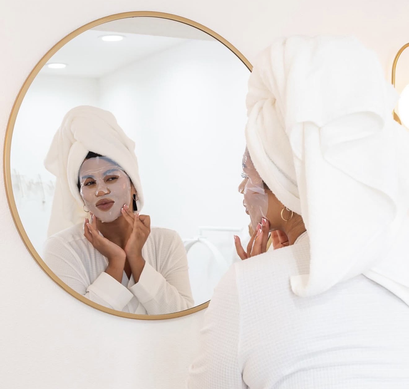 Person with towel on head looking at mirror, applying face mask, in a bright room.