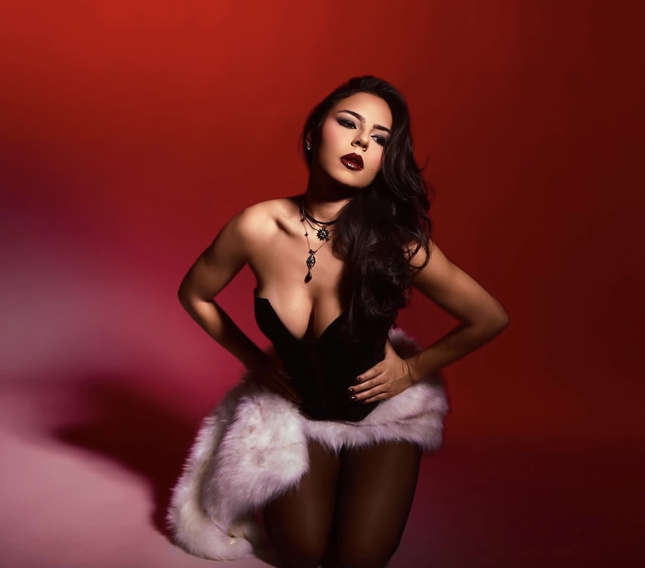 Woman with dark hair and makeup, wearing a black outfit with a white fur trim, posing against a red background.
