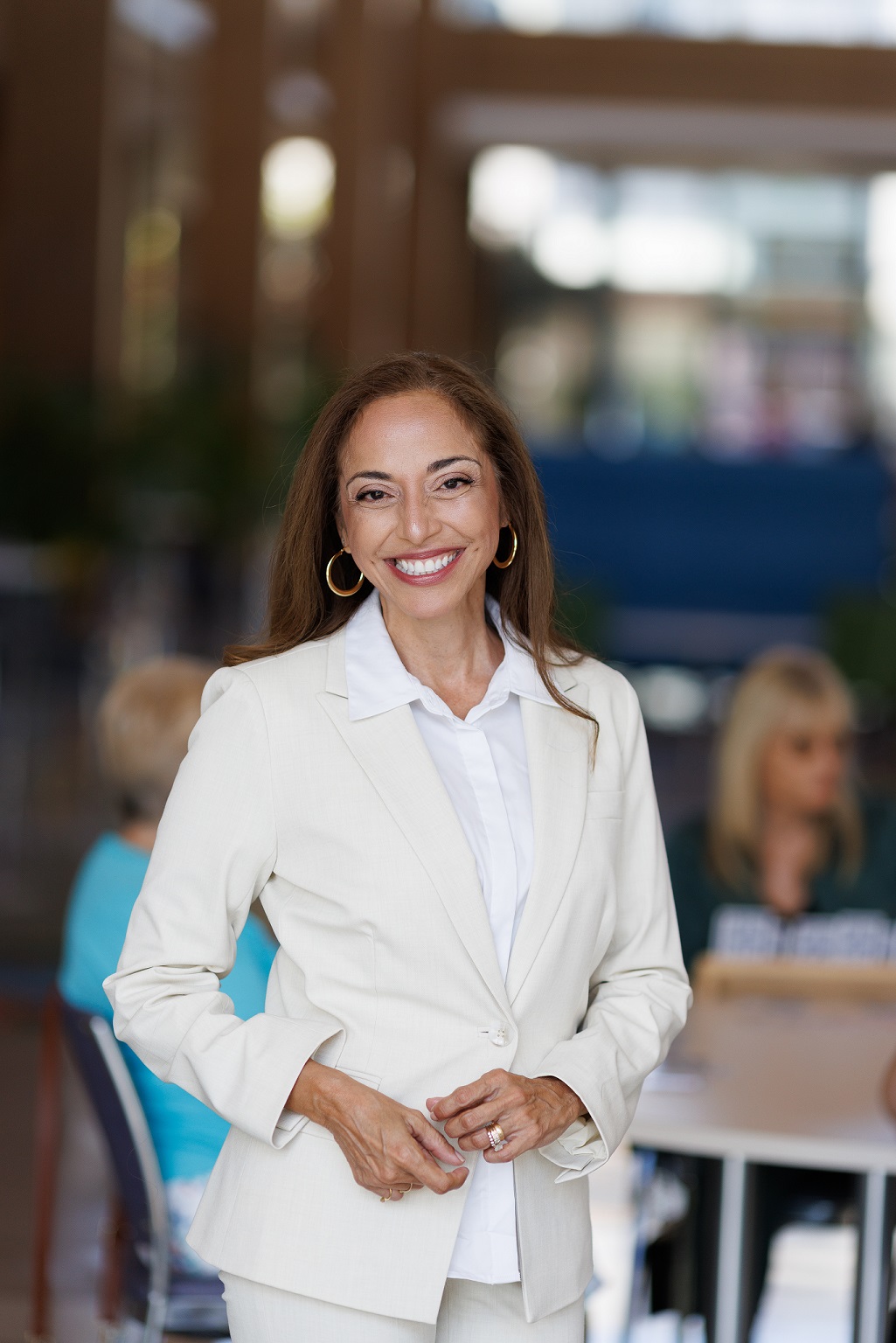 Smiling woman in white blazer and shirt standing indoors with blurred background of people and furniture.