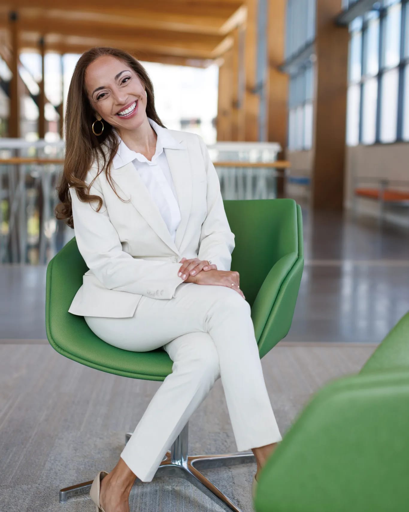 Smiling woman in white suit sitting in a green chair indoors with large windows and wooden beams.