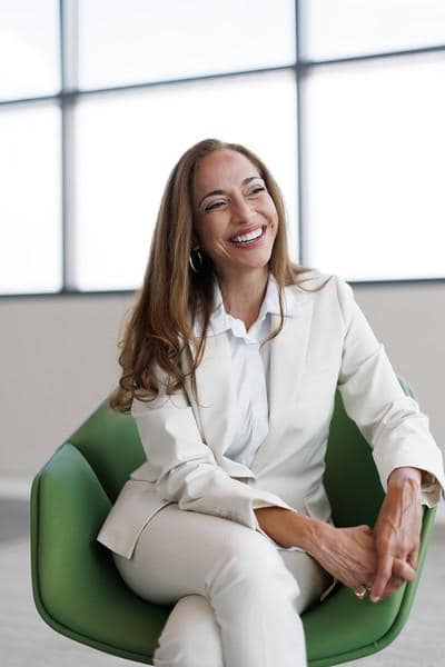 Woman with long hair smiling, sitting in a green chair in front of large windows, wearing a white blazer and shirt.