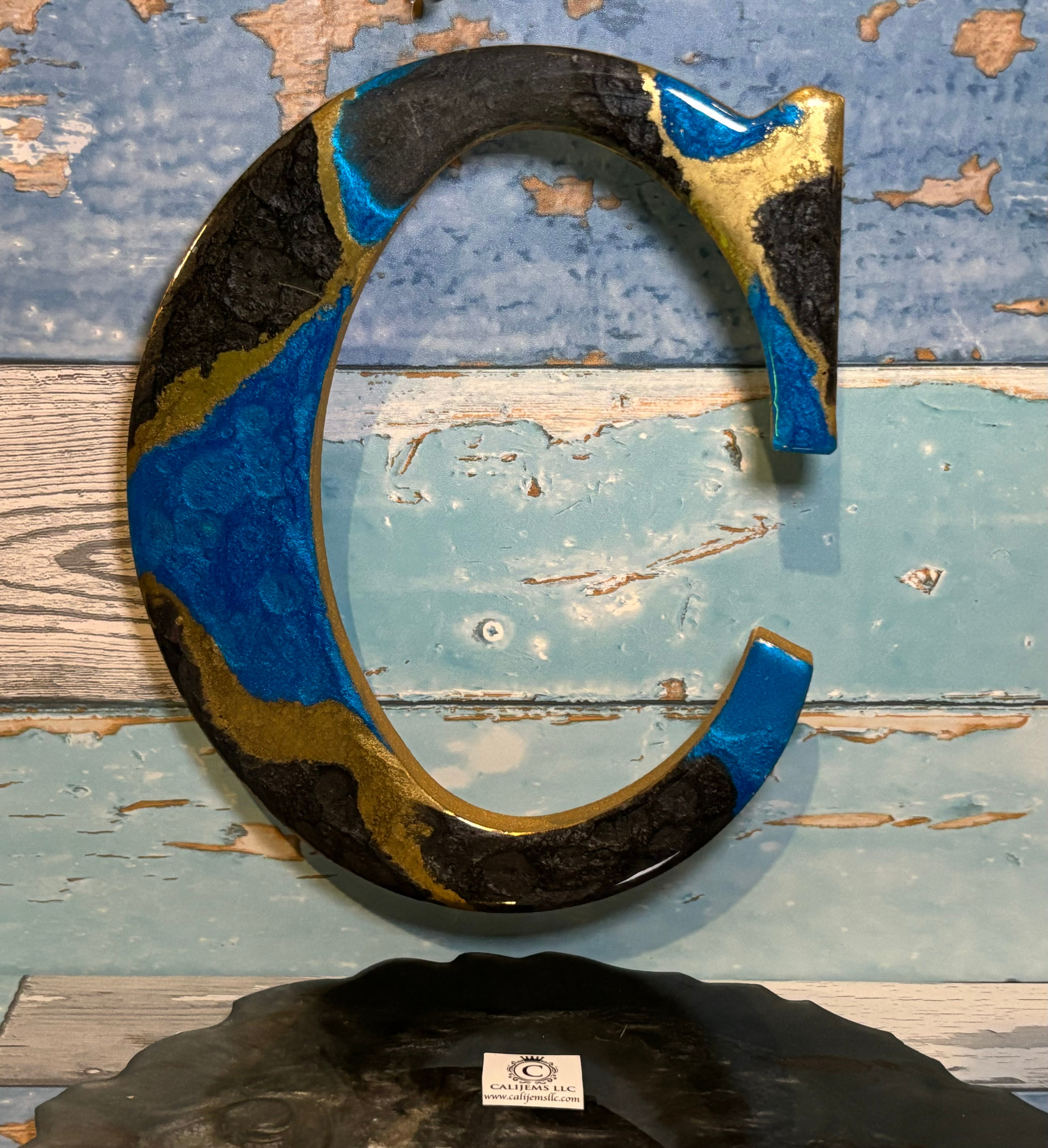 A large, colorful letter C sculpture with blue, black, and gold colors, mounted on a wooden surface with a weathered background.