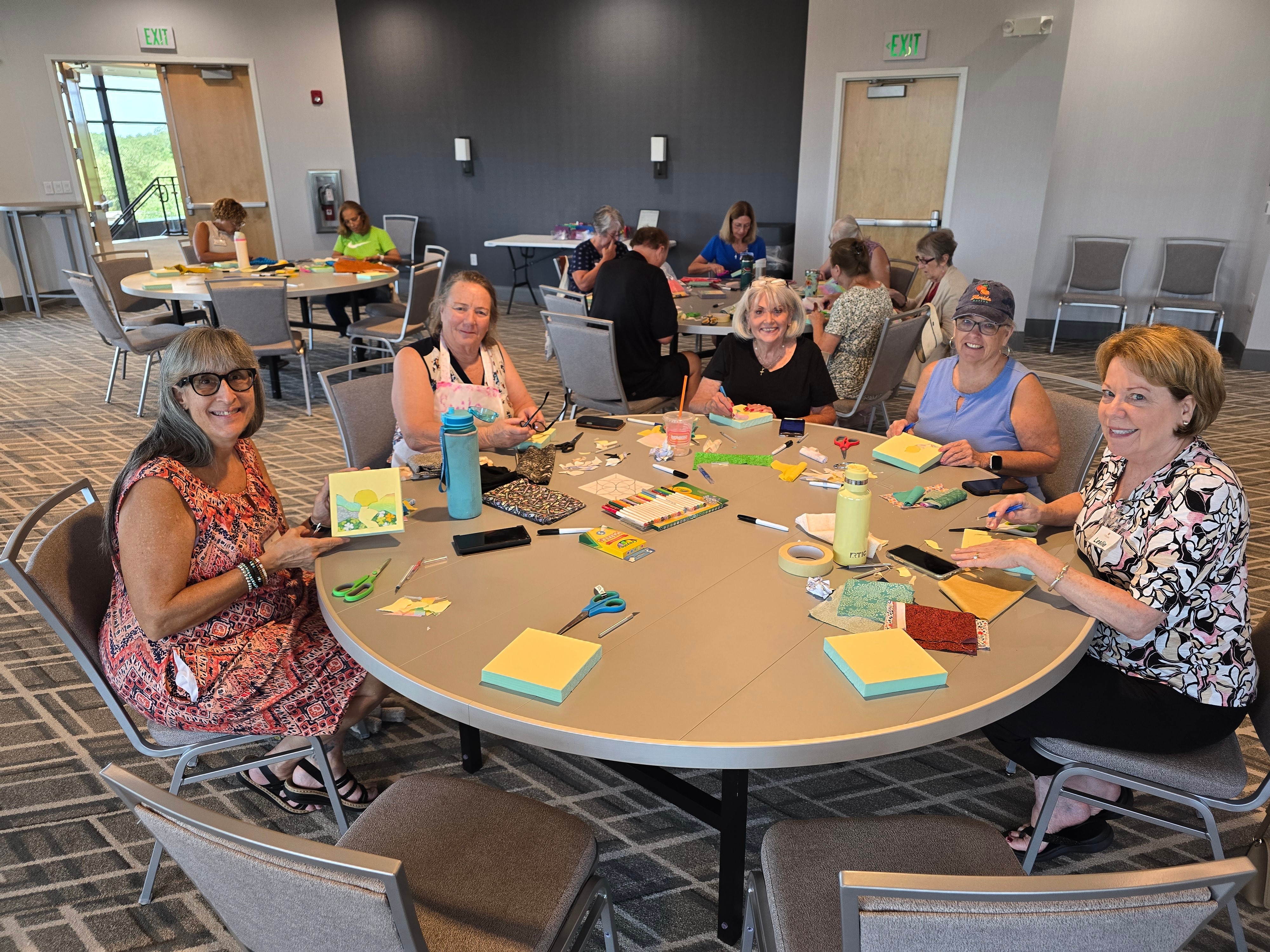 Group of women sitting around a large round table with colorful sticky notes and water bottles in a room with gray walls and large windows.