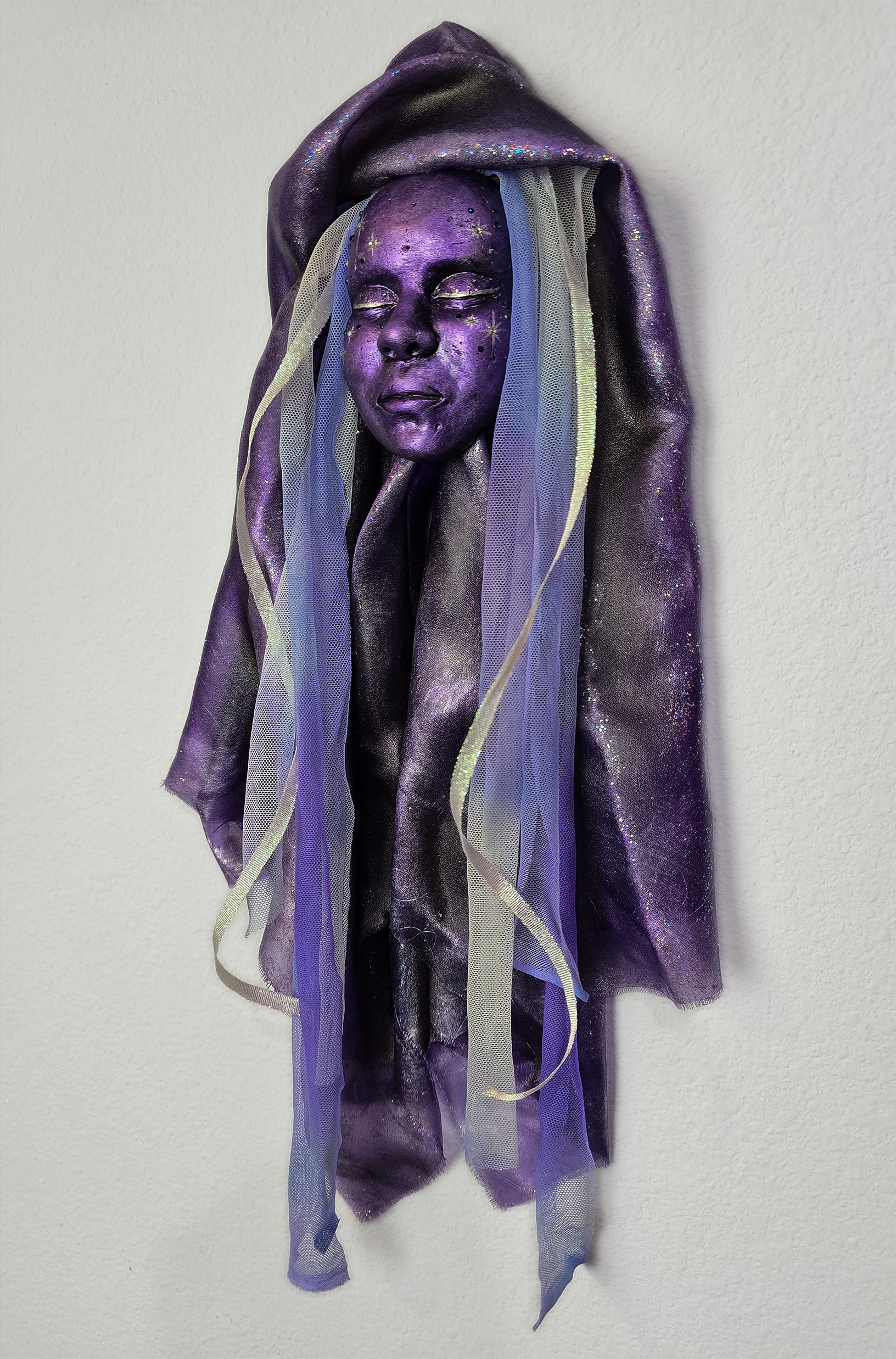 Sculpture of a woman's face with long, flowing hair and ribbons, painted in purple and blue tones, hanging against a plain wall.
