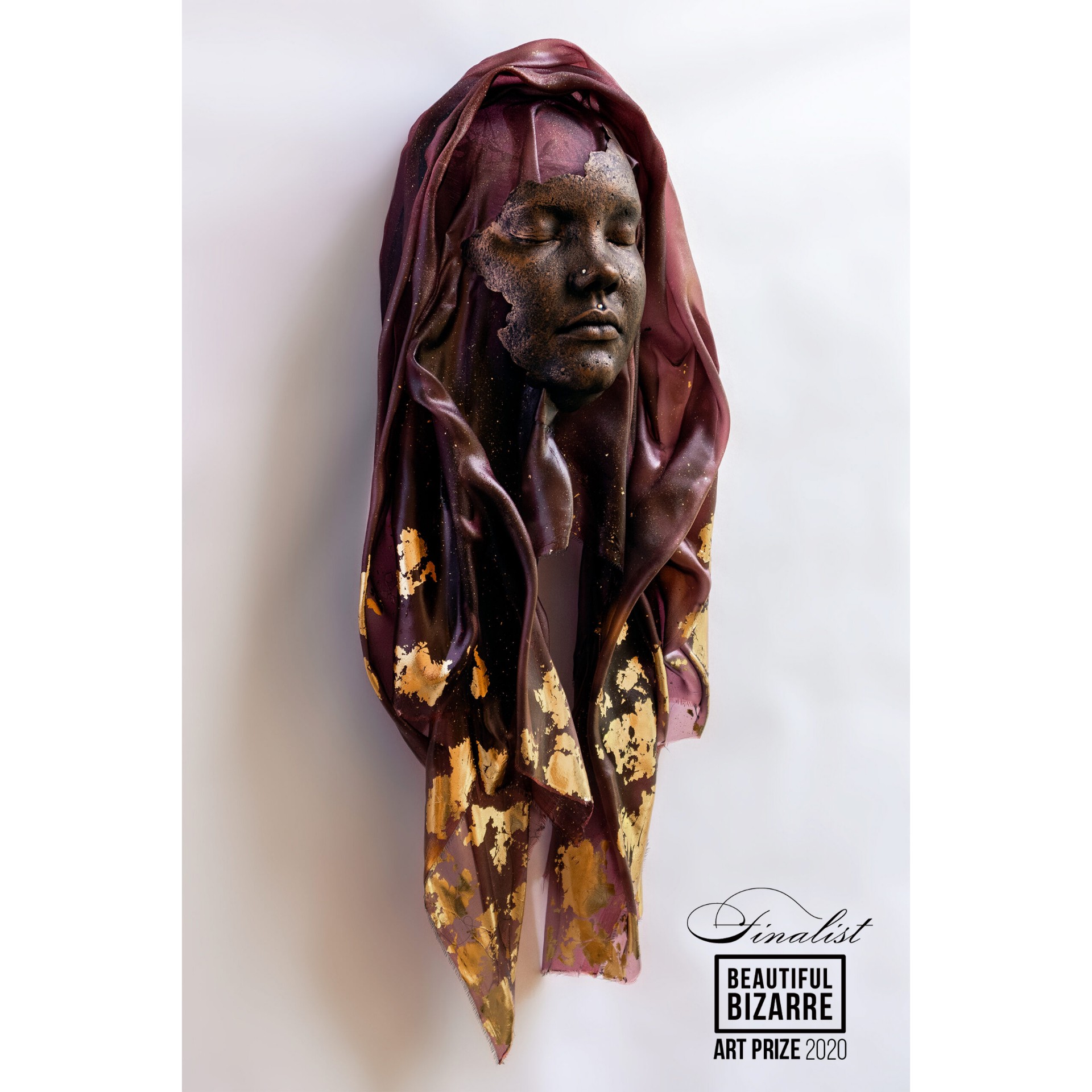 Sculpture of a woman's face with a maroon and gold fabric draped over her head and shoulders, mounted on a wall.