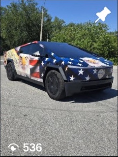 Car with a patriotic star and stripe design parked on a street, trees in the background, sunny day.