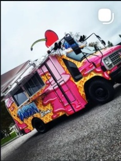 Colorful food truck with pink, yellow, and blue designs, parked on a street under a cloudy sky.