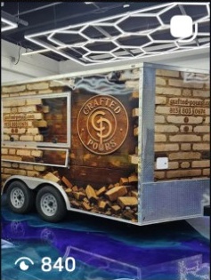 Food truck with brick wall design and logo, parked indoors with ceiling lights, trailer wheels visible, and blue floor lighting.