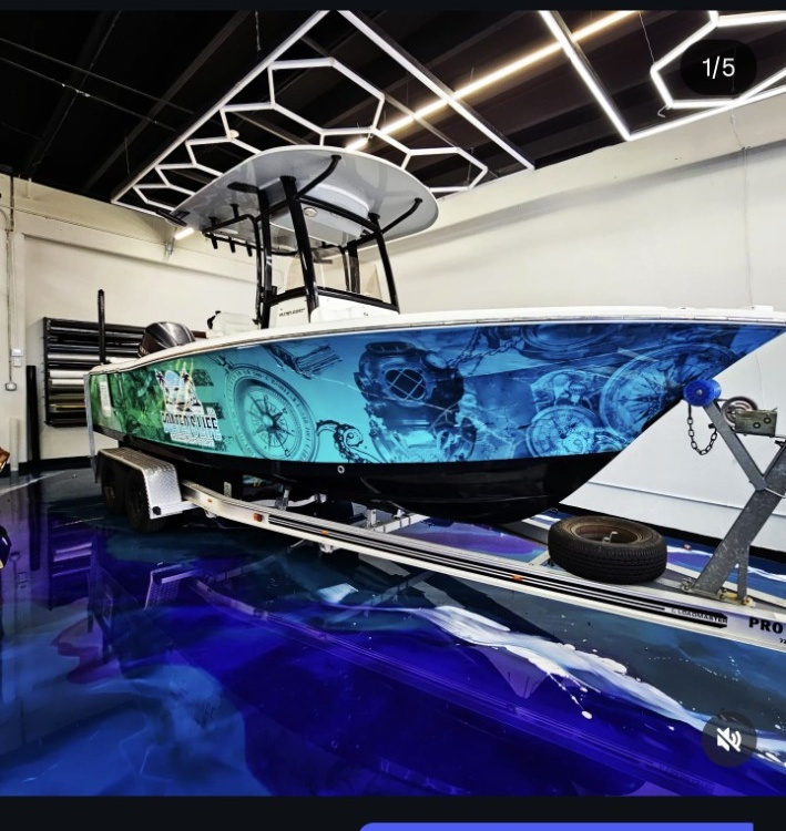 A boat on a trailer indoors with modern ceiling lights and a reflective floor, surrounded by equipment and a tire.