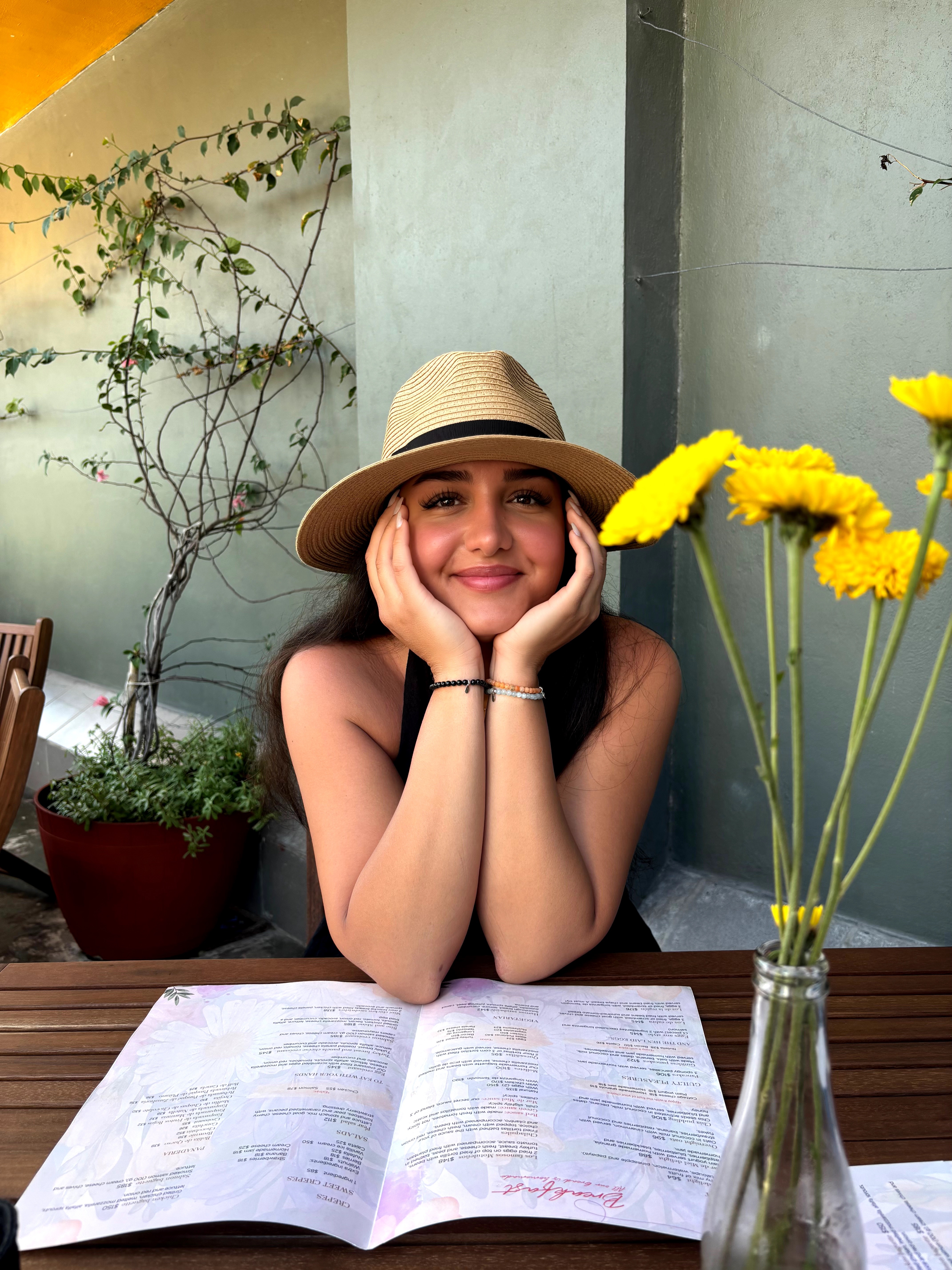Young woman with a hat smiling, resting her face on hands, sitting at a table with an open book, yellow flowers nearby.