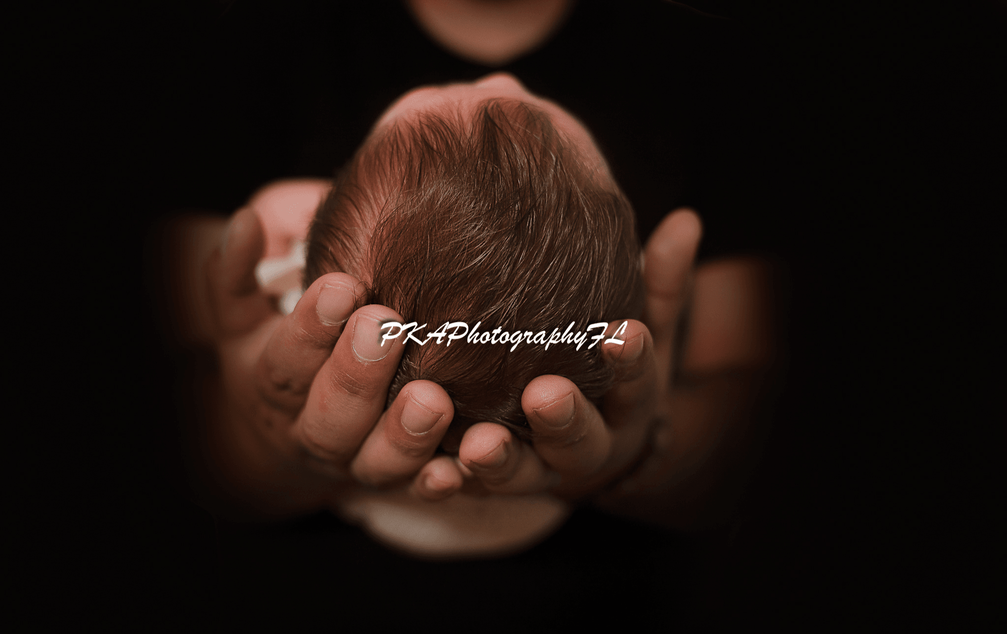 Person holding a small, curled-up baby with hands visible, dark background, soft lighting, and a watermark. Word count: 20.