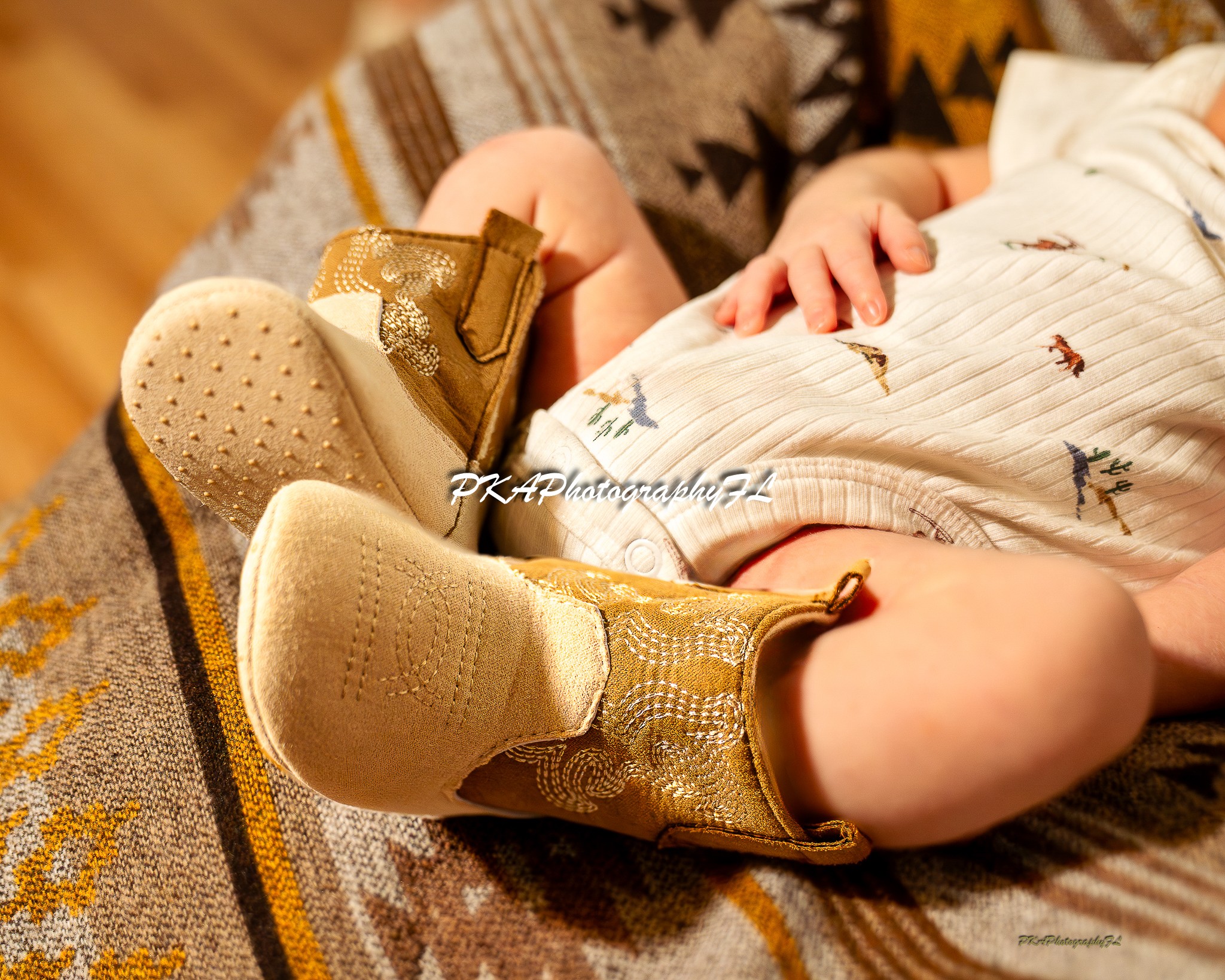 Baby wearing gold shoes, lying on a patterned blanket, with hands visible and wearing a striped outfit.