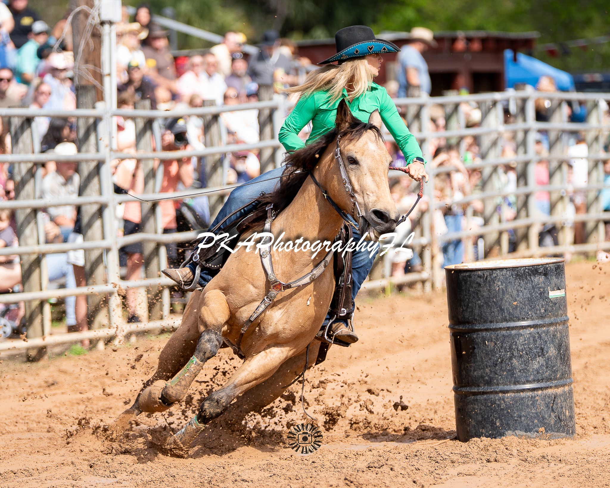 Female rider in green shirt and black hat riding a horse around a barrel during a rodeo event, crowd watching in background.