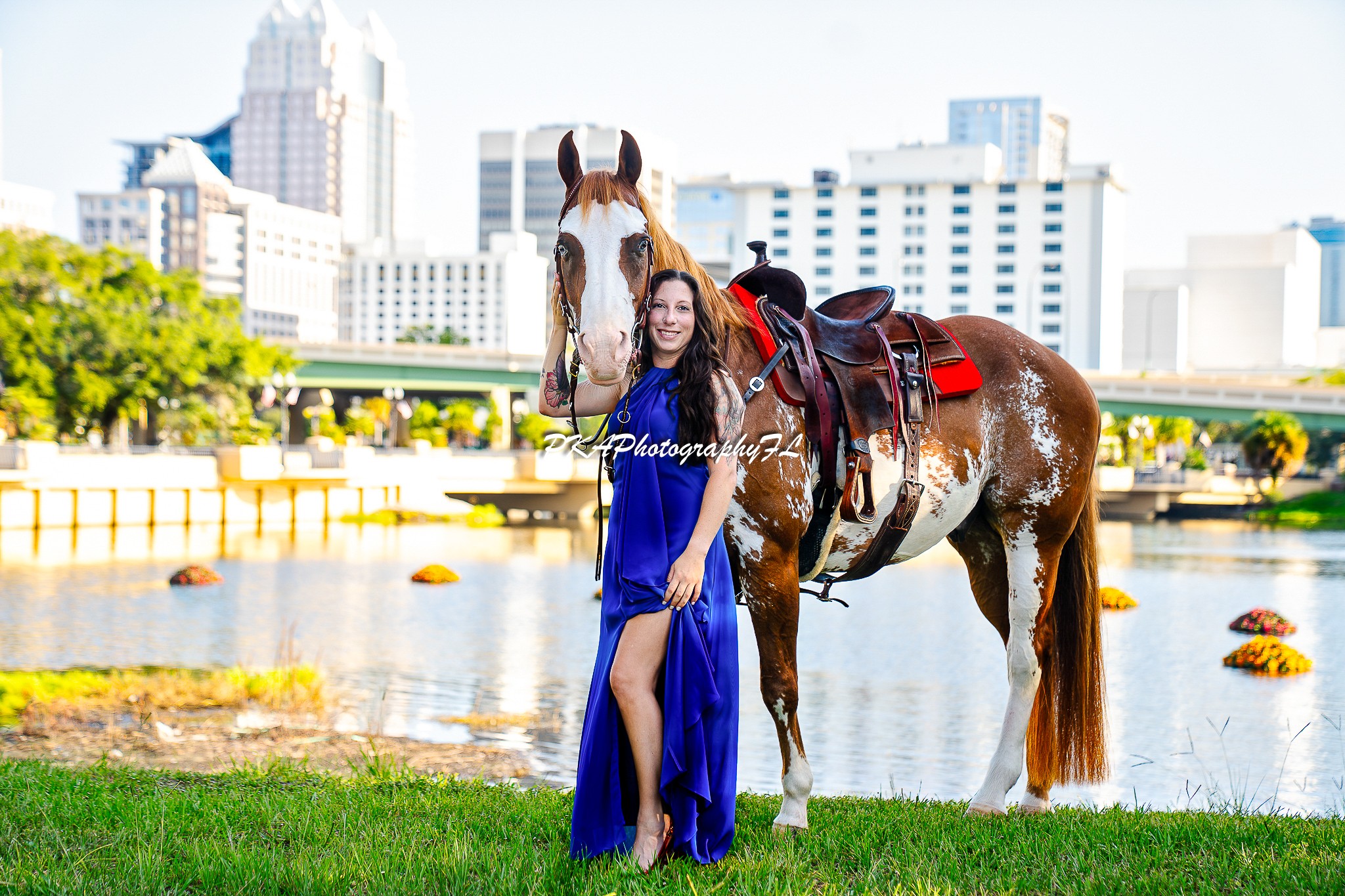 Woman in blue dress standing next to a horse near a river with city buildings in background.