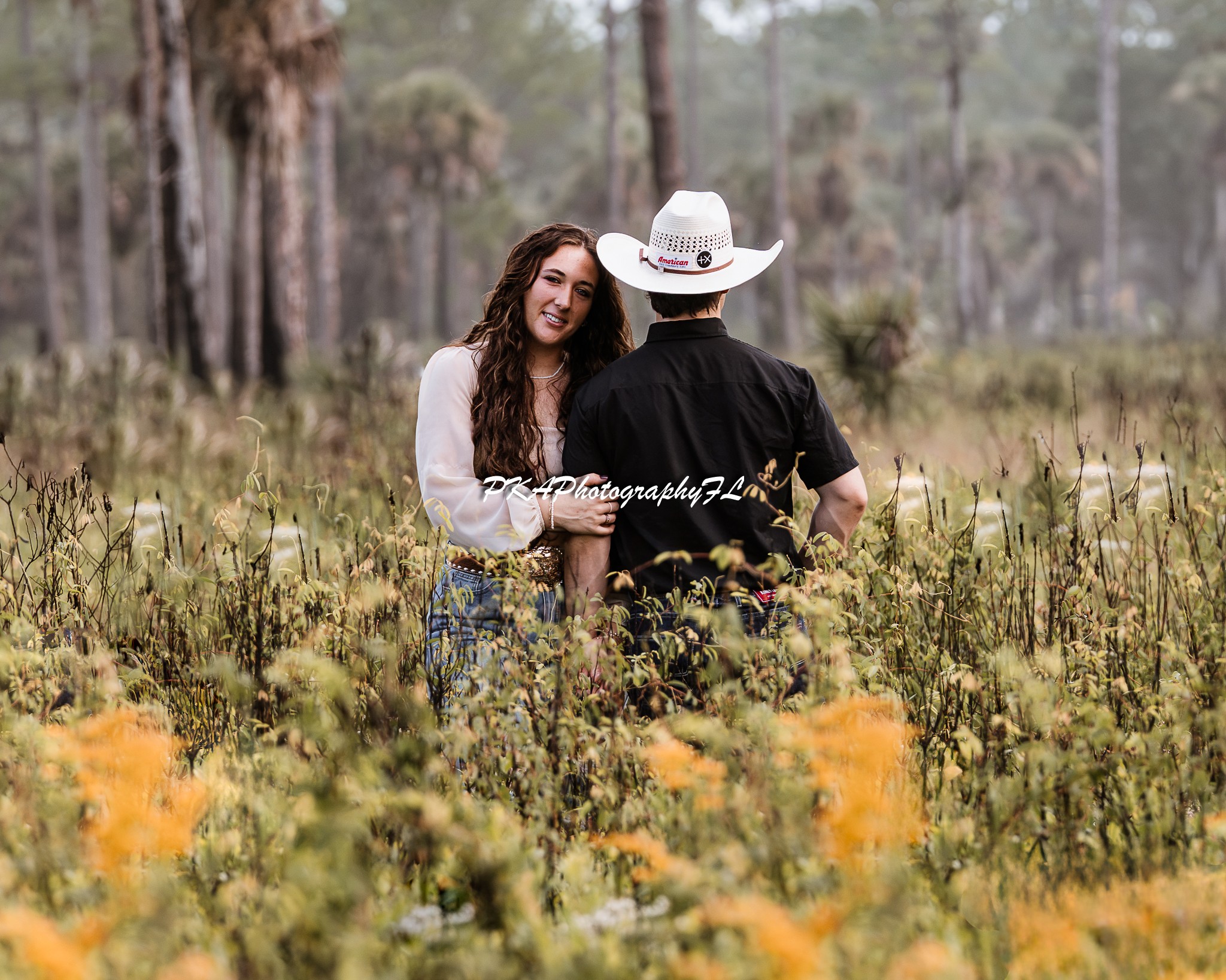 A woman and a person wearing a cowboy hat stand in a field with trees in the background, holding hands and talking.