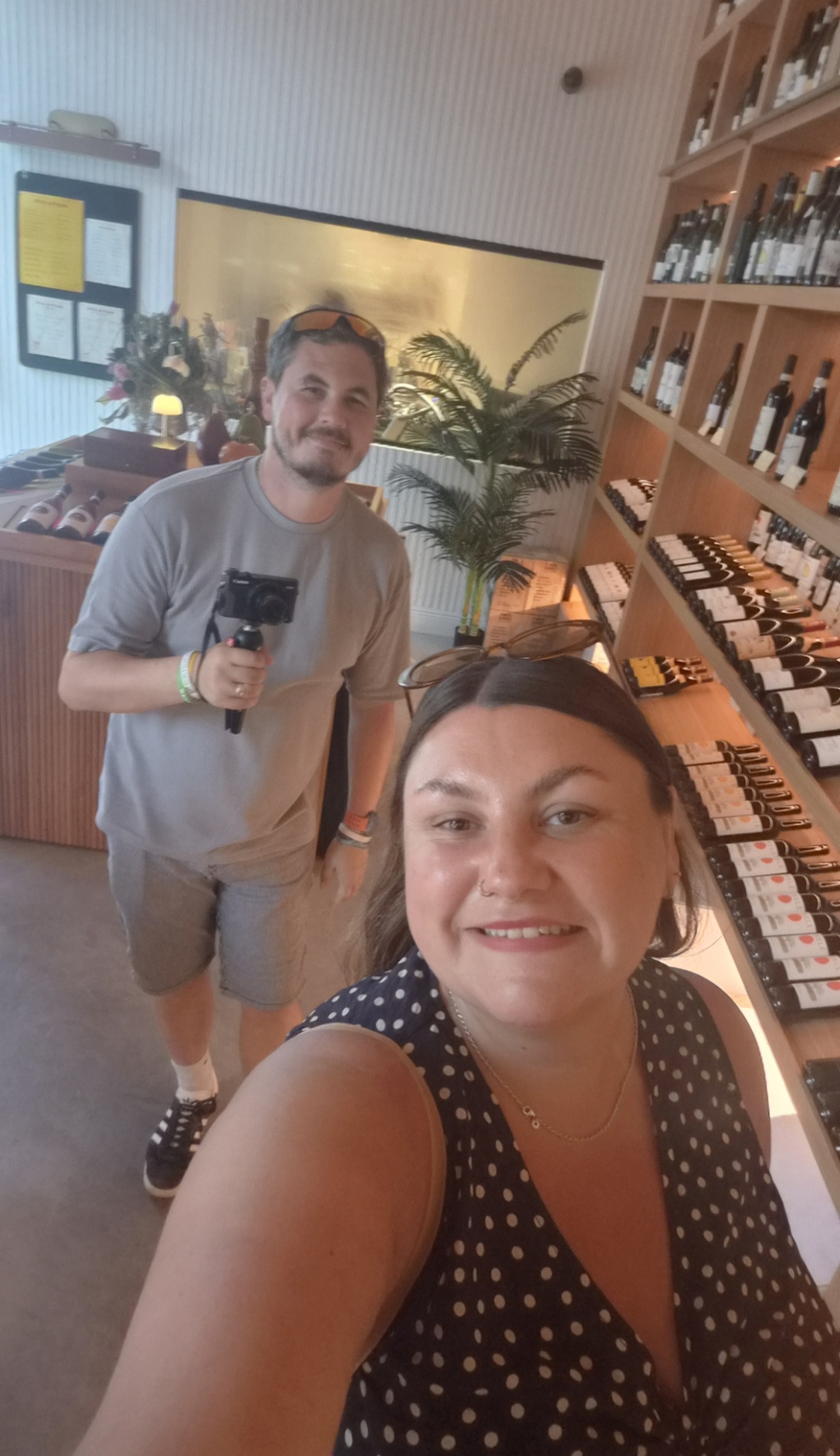Two people taking a selfie indoors, with shelves of products behind them, smiling at the camera.