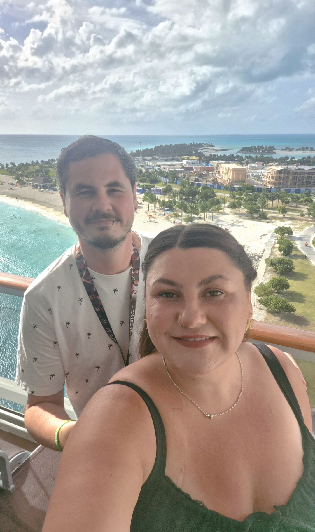 Two people taking a selfie on a balcony overlooking a beach and coastal area with buildings and greenery.