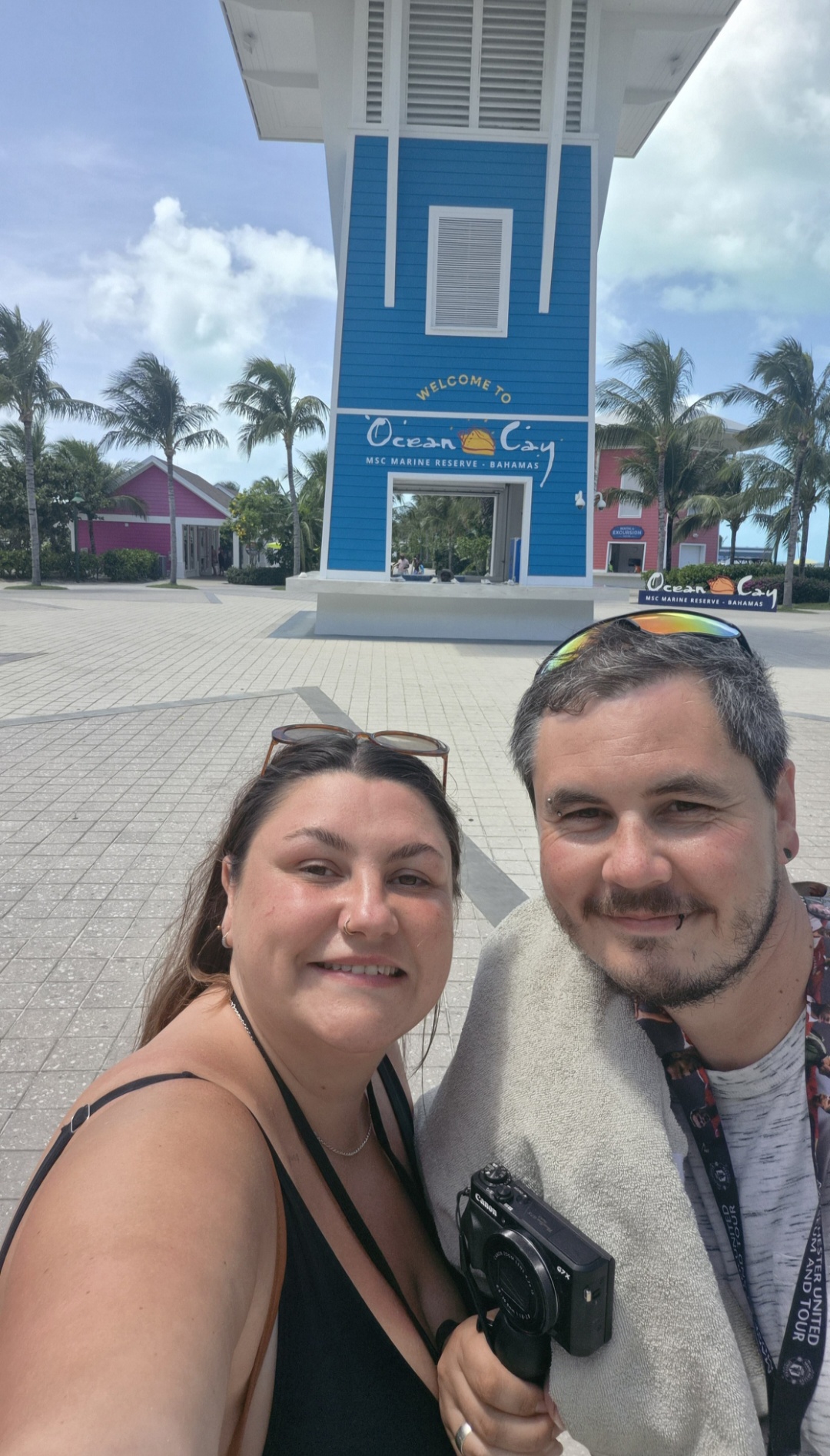Two people smiling in front of a large blue and white sign with palm trees and buildings in the background.