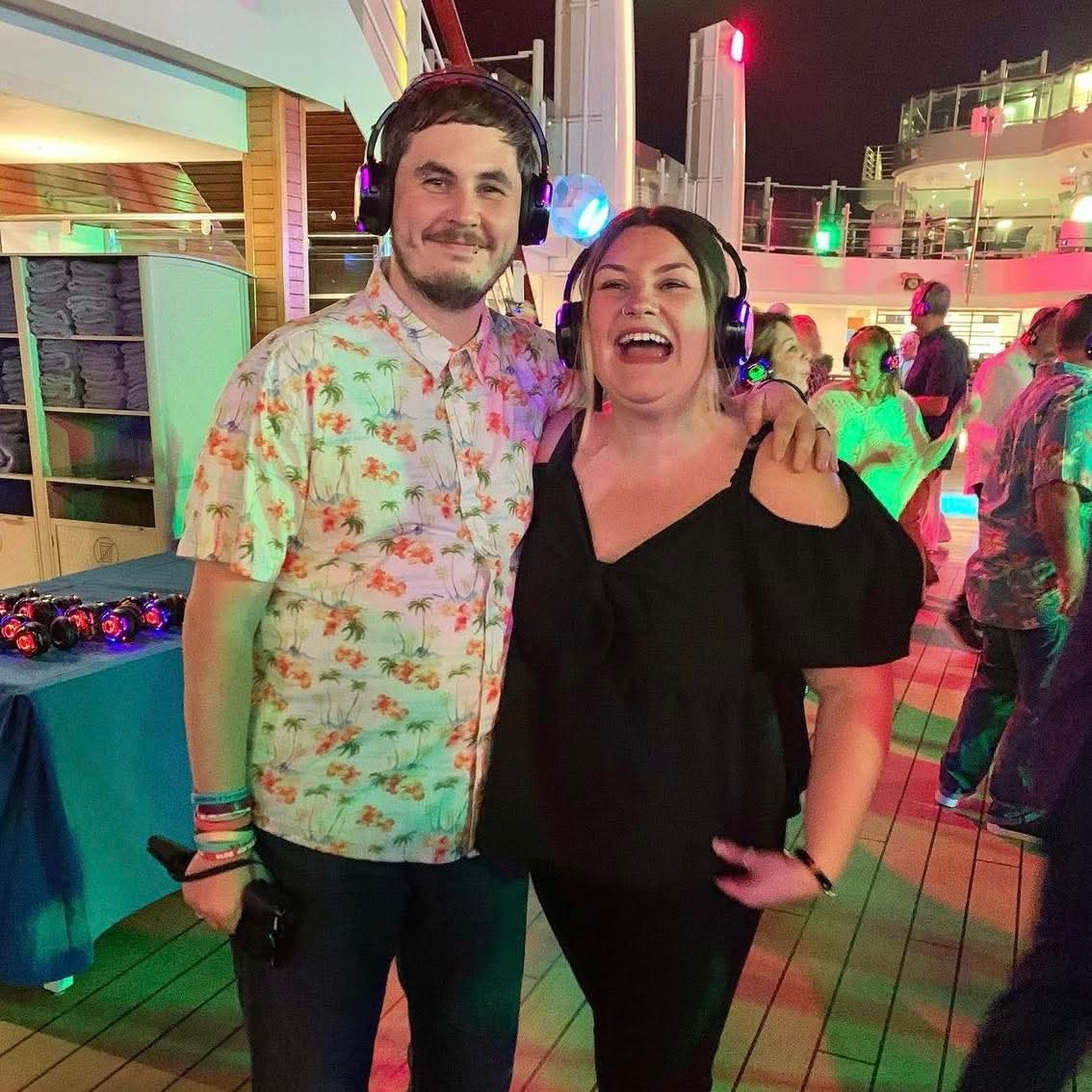 Two people smiling and wearing headphones, standing close together at a lively outdoor event at night.