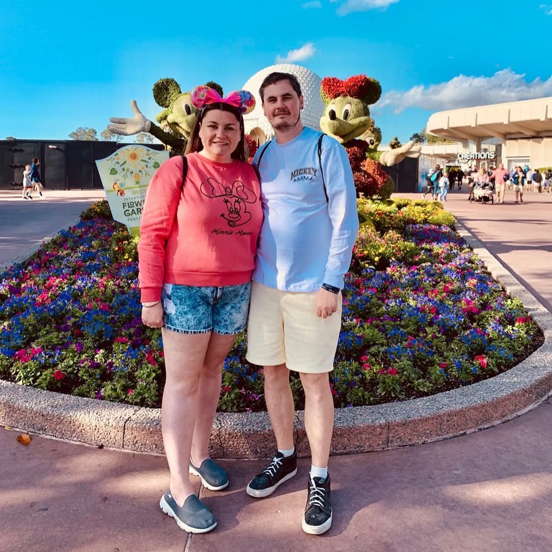 A smiling couple stands in front of a colorful flower bed with Disney character topiaries and a theme park entrance in the background.