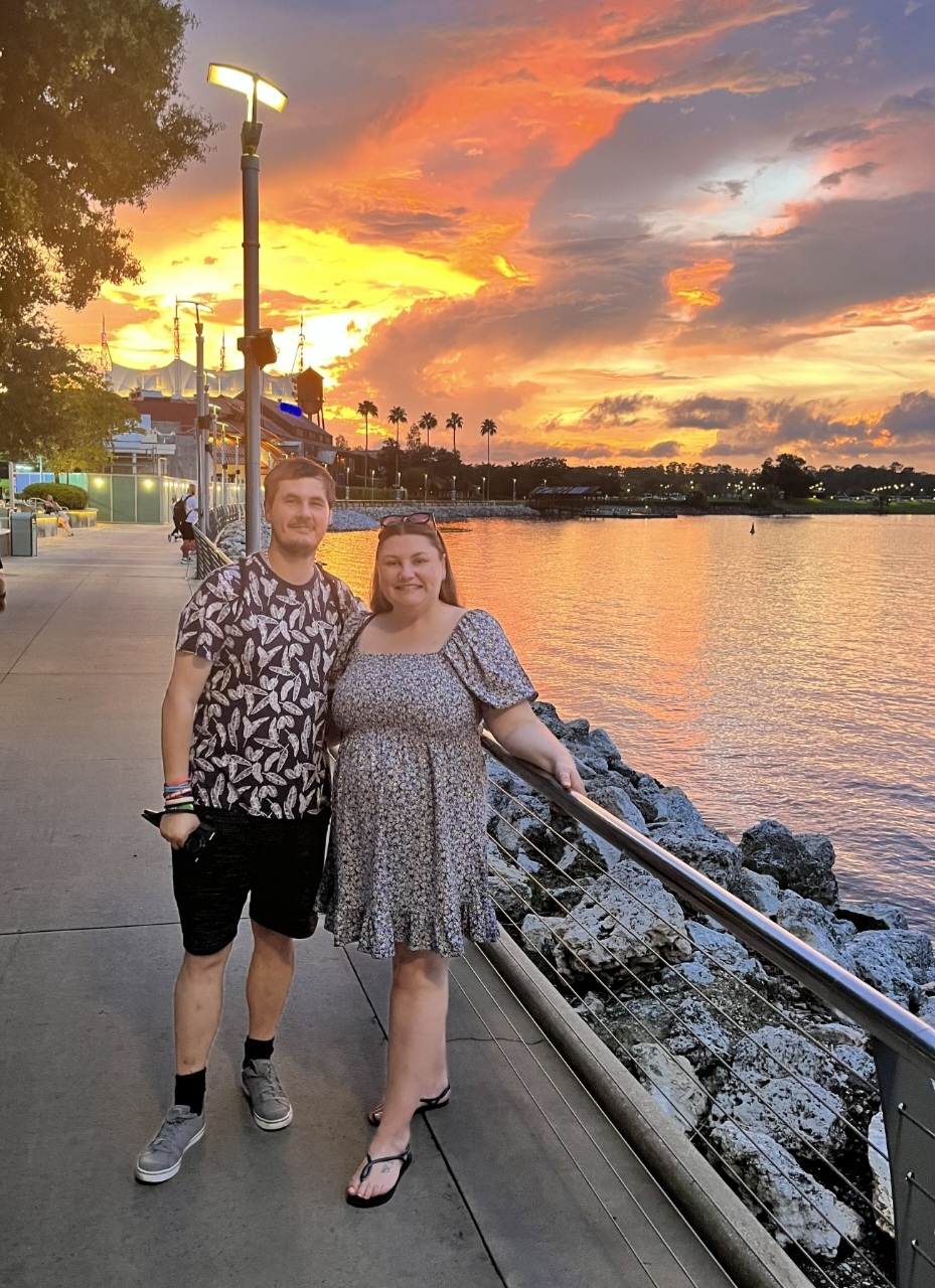 A man and woman stand on a waterfront promenade during sunset, with colorful sky and water behind them.