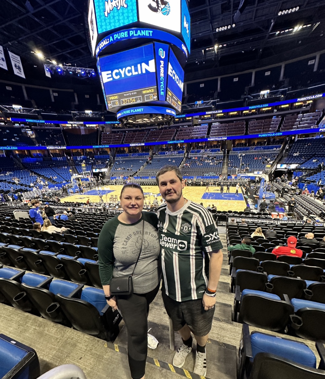 Two people standing in an arena with empty seats and a large digital scoreboard overhead.