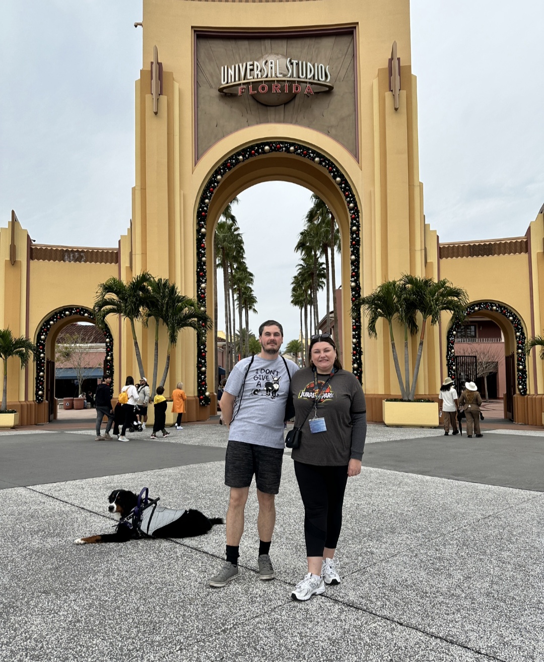 Two people stand in front of a large archway entrance with palm trees visible through it, at Universal Studios Florida, with a dog on a leash nearby.