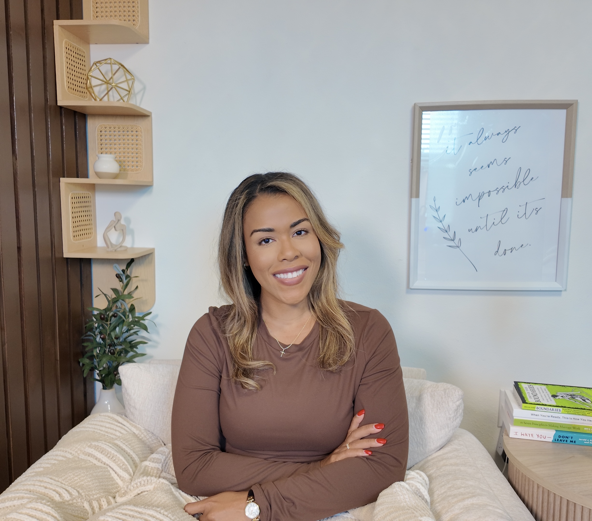 Woman with long hair smiling, sitting on bed with white pillows, in a room with wall art and shelves.