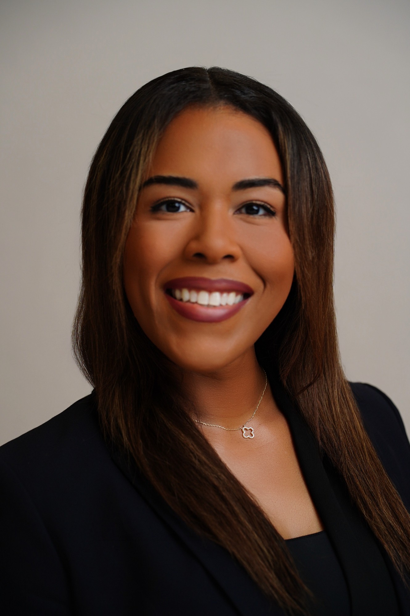 Portrait of a smiling woman with long dark hair, wearing a dark blazer and a necklace, against a plain background.