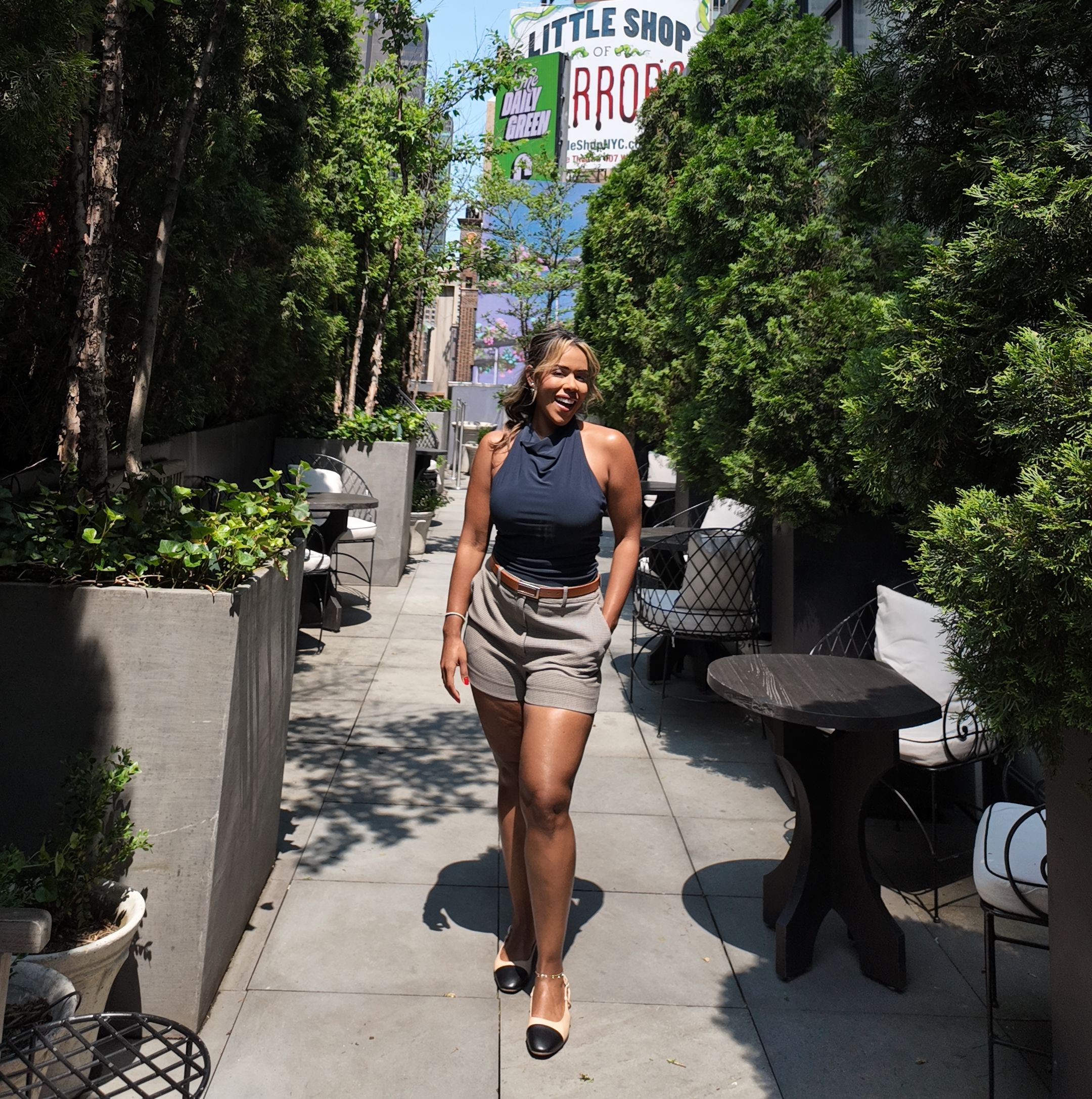 Woman in a dress standing outdoors on a sidewalk with trees and signs in the background.