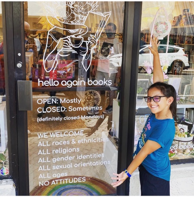 Girl with glasses and blue shirt smiling outside bookstore, waving at glass door with sign, books visible inside.