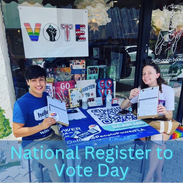 Two young people sitting at a table with voting materials, promoting National Register to Vote Day outside a store.