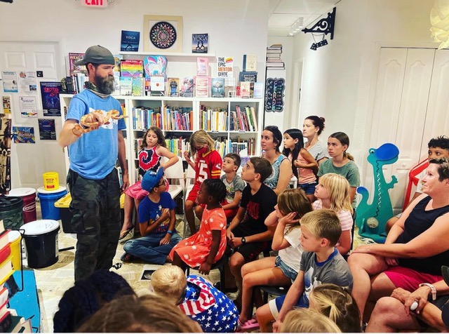 Man in blue shirt and hat holding a pizza slice, children seated and watching in a room with bookshelves and decorations.