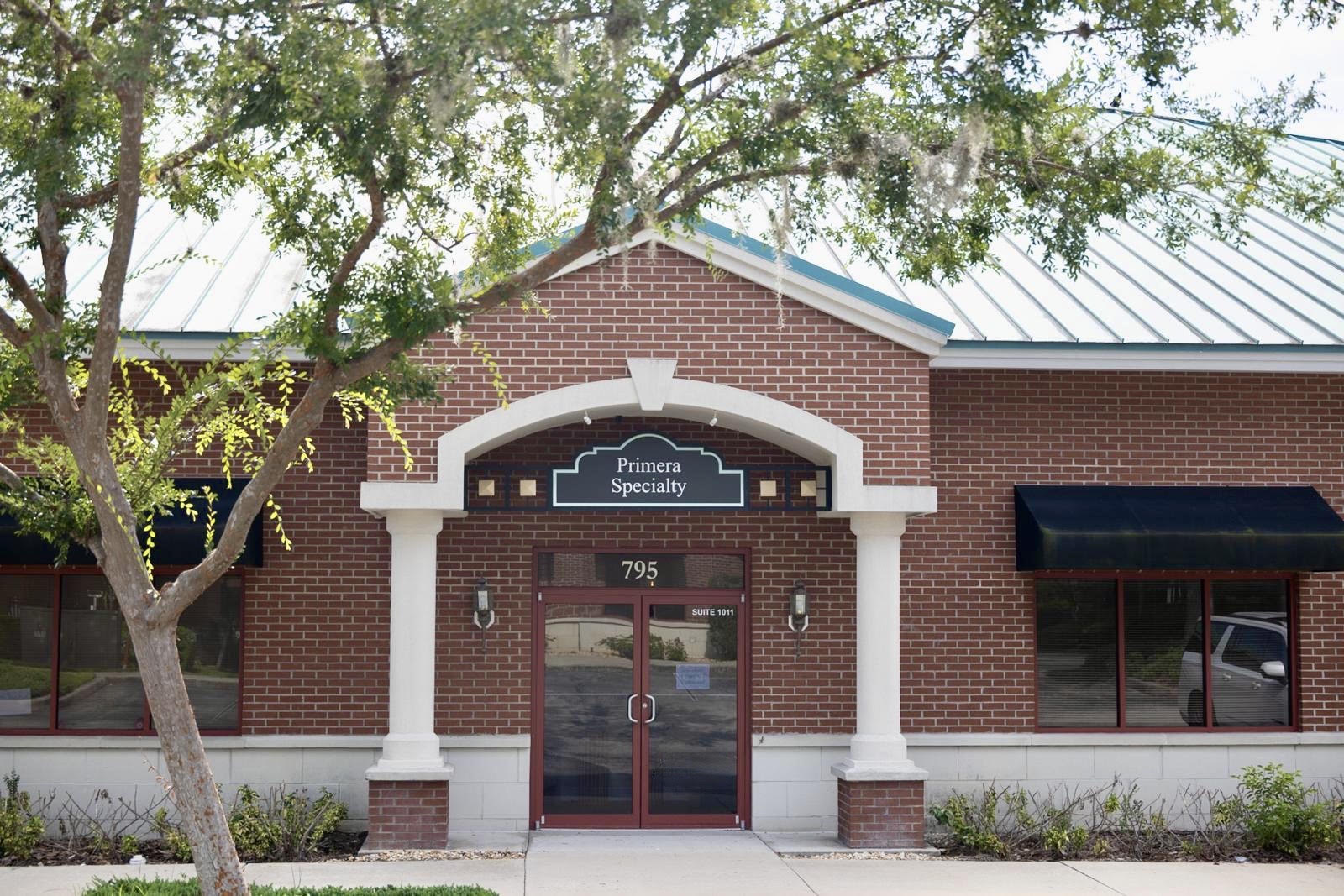 Brick building entrance with white archway, glass doors, and a sign reading 'Primary Specialty', surrounded by trees and windows.