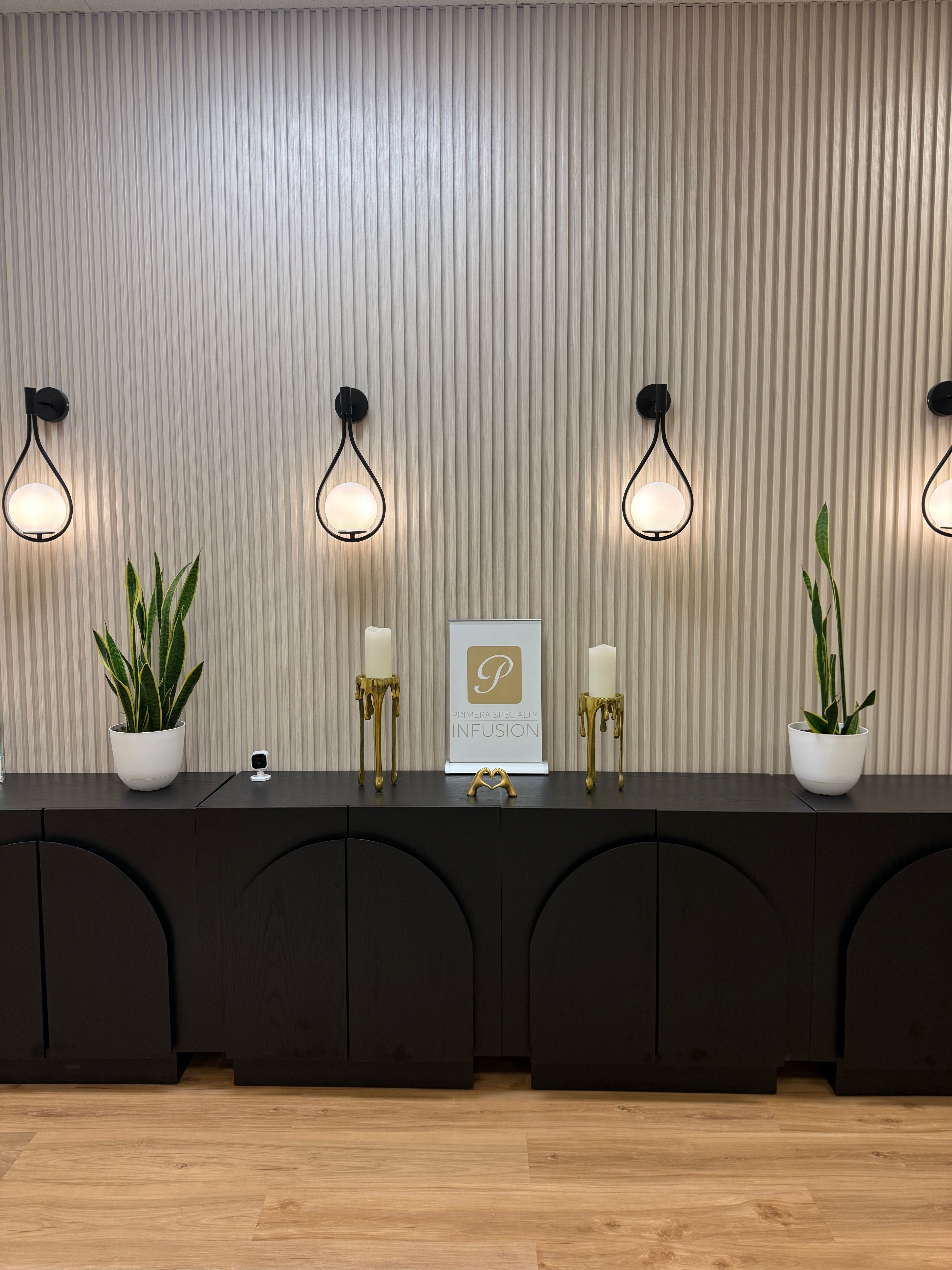 Interior with wall-mounted lights, potted plants, and decorative items on a shelf, against a striped wall and wooden floor.
