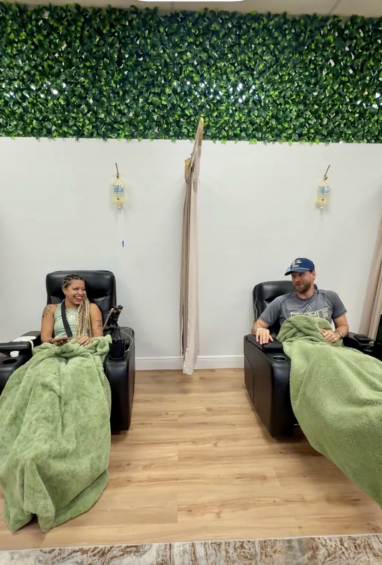 Two people sitting in chairs with green blankets, in a room with a white wall and green plant decor, wooden floor.