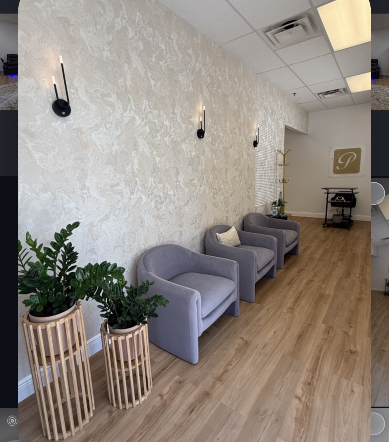 Waiting area with three gray armchairs, potted plant, and wall-mounted lights on textured wall, wooden floor, and ceiling lights.